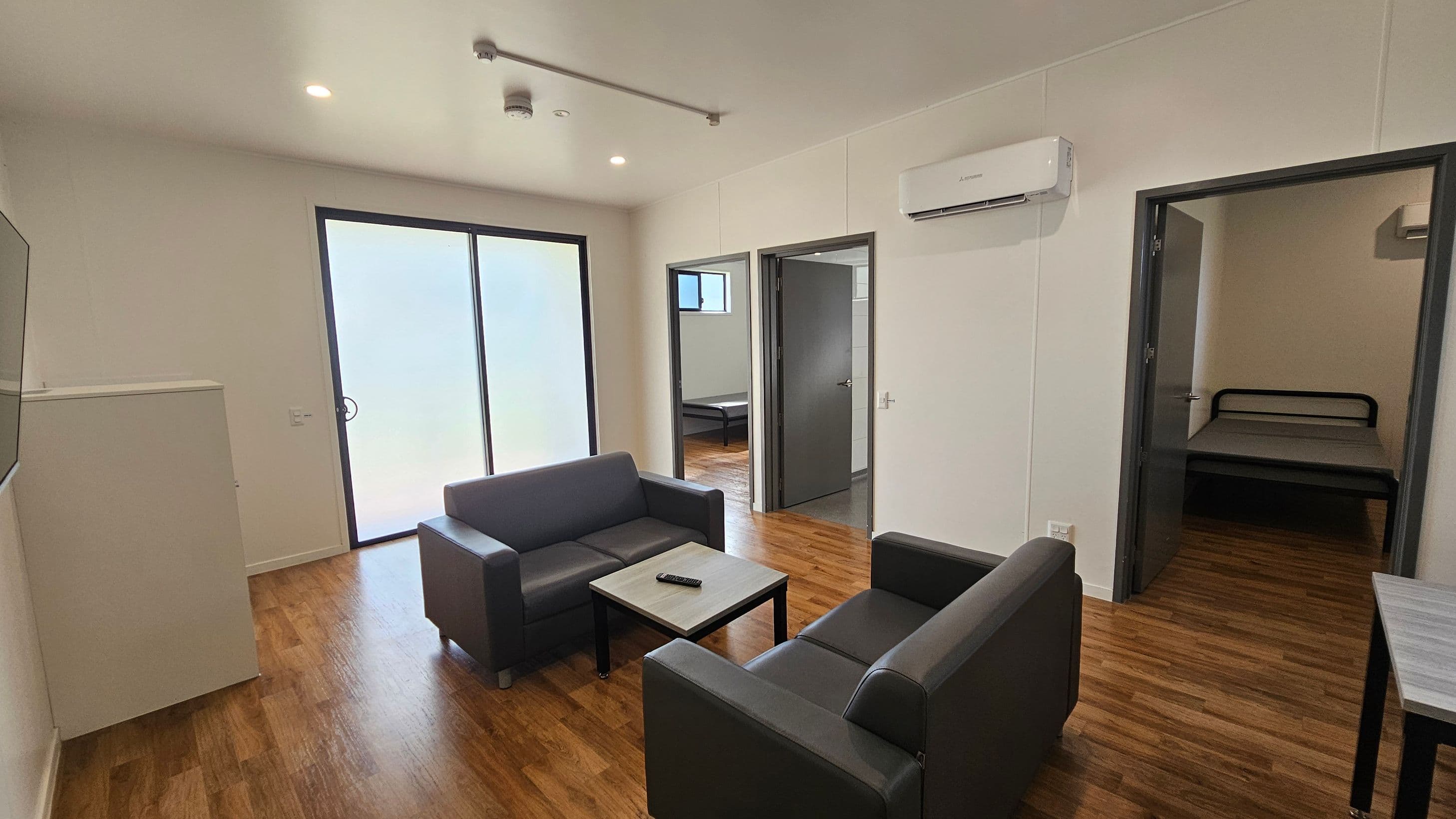 A modern living room with wooden flooring. In the centre, two dark grey sofas face each other with a small coffee table between them, holding a remote control. On the left, large sliding glass doors allow natural light in. On the right, two doorways lead to separate rooms, each containing a bed. An air conditioning unit is mounted on the wall above the right doorway. The ceiling is white with recessed lighting.