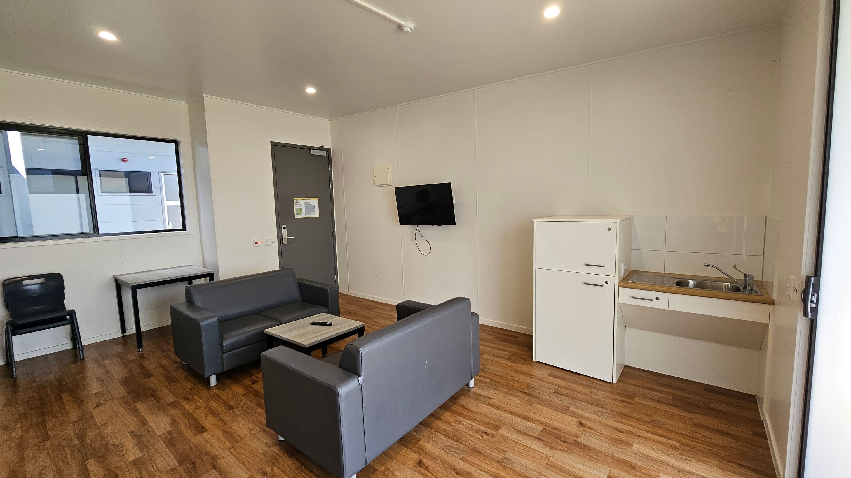 On the left, there is a dark-coloured chair next to a small table with a light wood surface. A large window with a black frame allows light into the space. To the right, a grey door leads to another area, and a television is mounted on the wall next to it. A white fridge stands beside a compact kitchen area with a sink and a countertop. The floor is covered in light wooden laminate, providing a warm tone. The ceiling is flat and painted white, with several recessed lights.