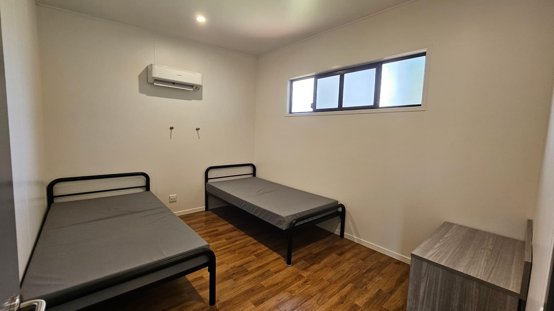On the left, a single bed with a black metal frame is positioned against the wall, covered with a grey mattress. To the right, another similar bed is set parallel, also with a grey mattress. An air conditioning unit is mounted above the beds, and two wall hooks provide additional storage. In the background, a narrow window with multiple panes allows light into the room. The floor is covered in a light wooden laminate, and the ceiling is flat with bright recessed lights.
