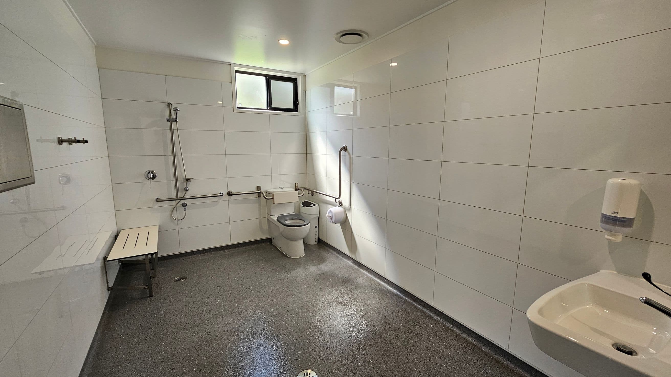 A spacious, accessible bathroom with white tiled walls and a grey speckled floor. On the left, there's a shower area with a bench and grab bars. In the centre, a toilet with additional grab bars and a small window above. On the right, a sink with a soap dispenser mounted on the wall. The ceiling is white with recessed lighting.