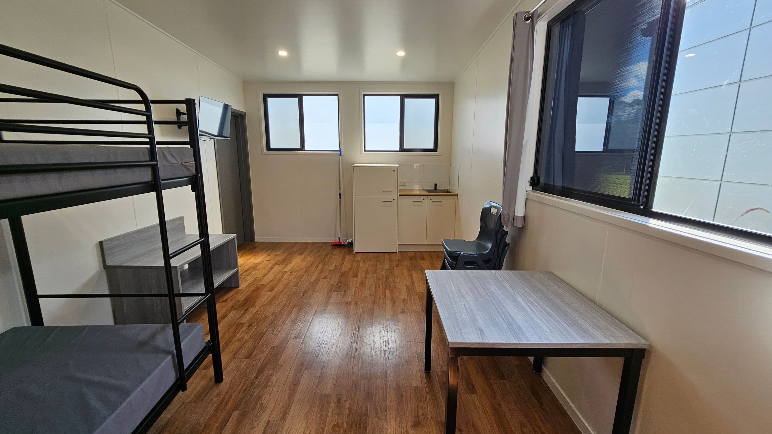 On the left, a black metal bunk bed occupies the corner, with a small grey bedside table beside it. To the right, a grey plastic chair sits adjacent to a light wood table. A compact kitchenette is positioned against the wall, featuring a sink and storage cabinet below. In the background, two rectangular windows allow natural light into the room, partially covered by light curtains. The floor is finished in a light wooden laminate, and the ceiling is flat and white, fitted with recessed light.