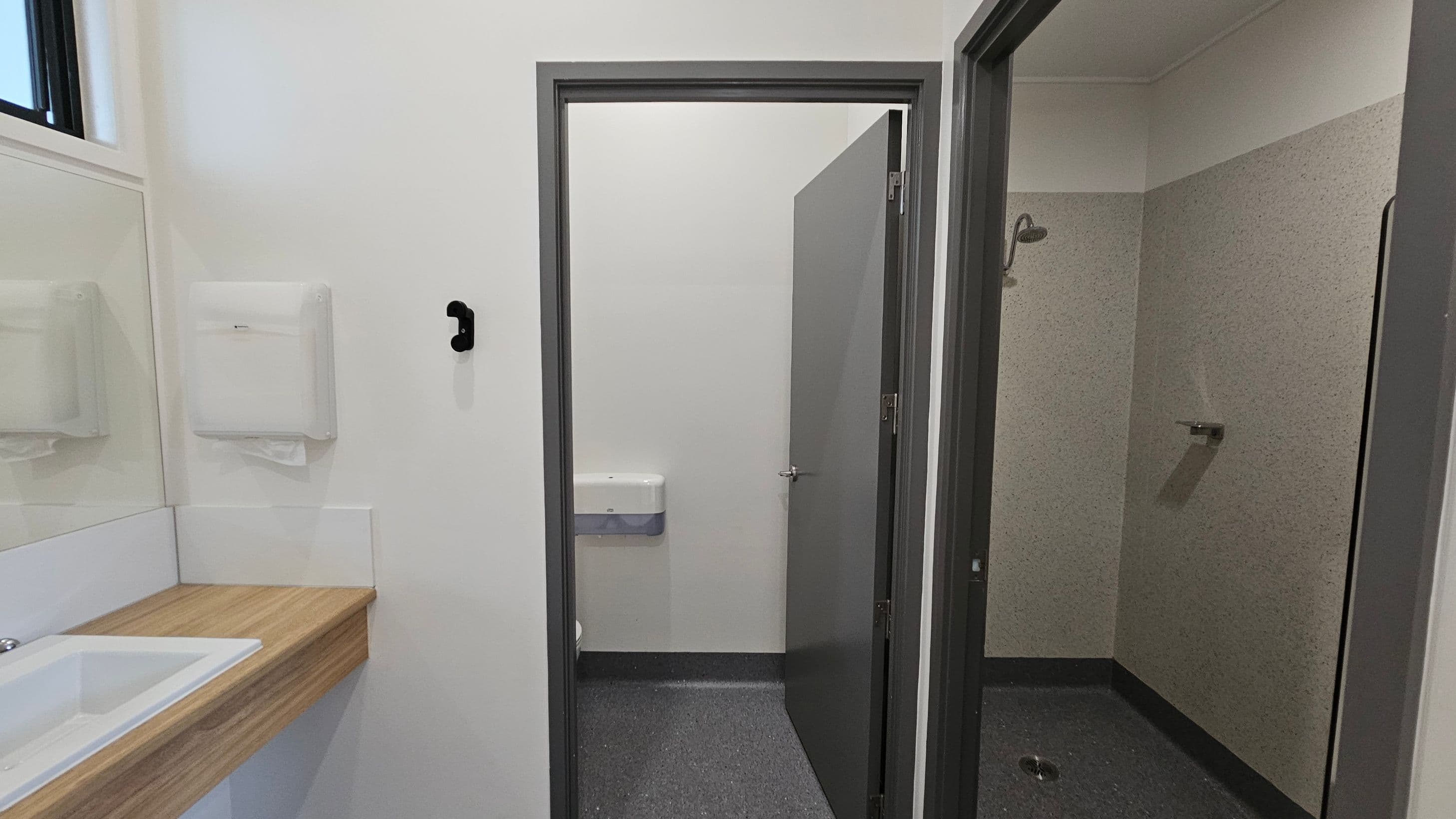 To the left, a wooden countertop with a white sink is located beneath a large mirror, with a paper towel dispenser mounted on the wall above it. There is a black hook on the wall nearby. On the right, an open grey door leads to a shower area, which features a wall-mounted showerhead and a small drain on the floor. A second door, also grey, is slightly ajar, revealing a toilet with a flush mechanism visible. The floor is a dark grey, textured surface, and the ceiling is flat and white.
