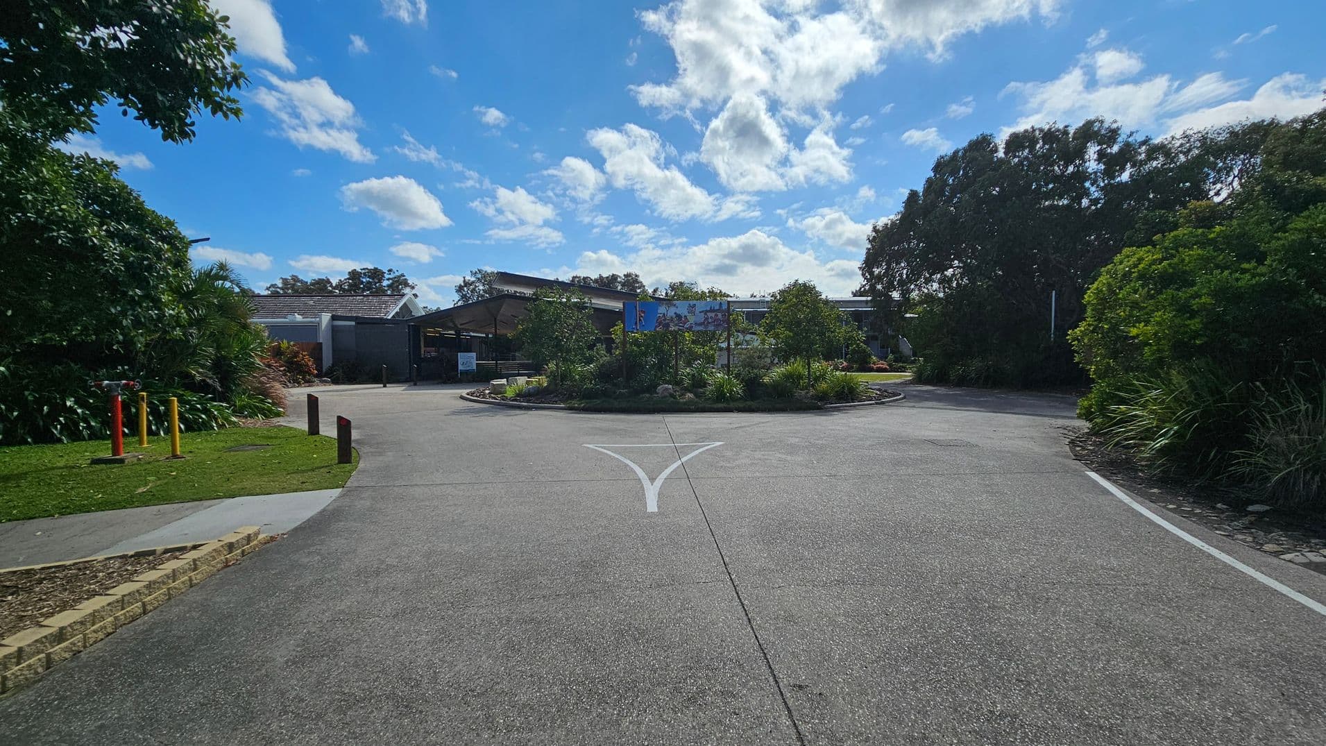 A paved road leads to a roundabout with lush greenery and small trees in the centre. On the left, there are colourful bollards and a grassy area with dense foliage. On the right, more trees and bushes line the road. In the background, a building with a sloped roof and a large sign is visible, partially obscured by the vegetation. The sky is filled with scattered clouds.