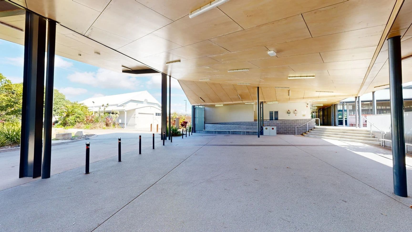 The area features a smooth concrete floor, extending underneath a canopy with a wooden panel ceiling supported by black metal columns. On the left, lush greenery and shrubs are visible beyond a row of low black bollards. In the background, a building with a peaked roof is partly visible. To the right, a set of steps leads up to a higher level, with a low wall behind it. There are several benches against the wall, and wall-mounted features can be seen as well.