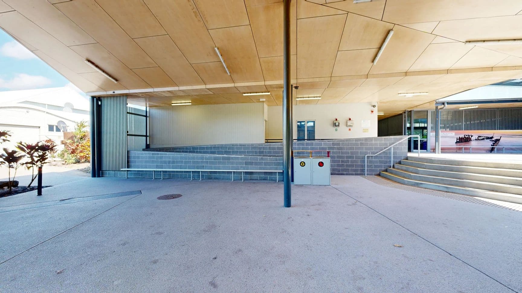 The area features a smooth, light grey concrete floor extending across the open space. On the left, a row of low shrubs adds greenery beside a vertical panel of grey cladding, while on the right, there are several steps leading up to a raised platform. The back wall has a panel with windows and two fire safety equipment units mounted near a white wall. The ceiling above is constructed of wooden panels, fitted with overhead lights , supported by tall, black metal columns in the foreground.