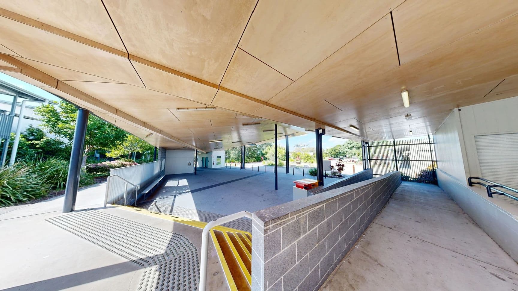 The area features a smooth concrete floor with a slight gradient leading towards a loading ramp on the left side, where a textured yellow edge marks the transition. On the right, a wall of grey bricks rises beside a metal railing, leading to a more enclosed space. The ceiling is wooden, with panels and overhead lights positioned evenly along its surface, supported by black metallic columns. In the background, greenery is visible through glass doors.
