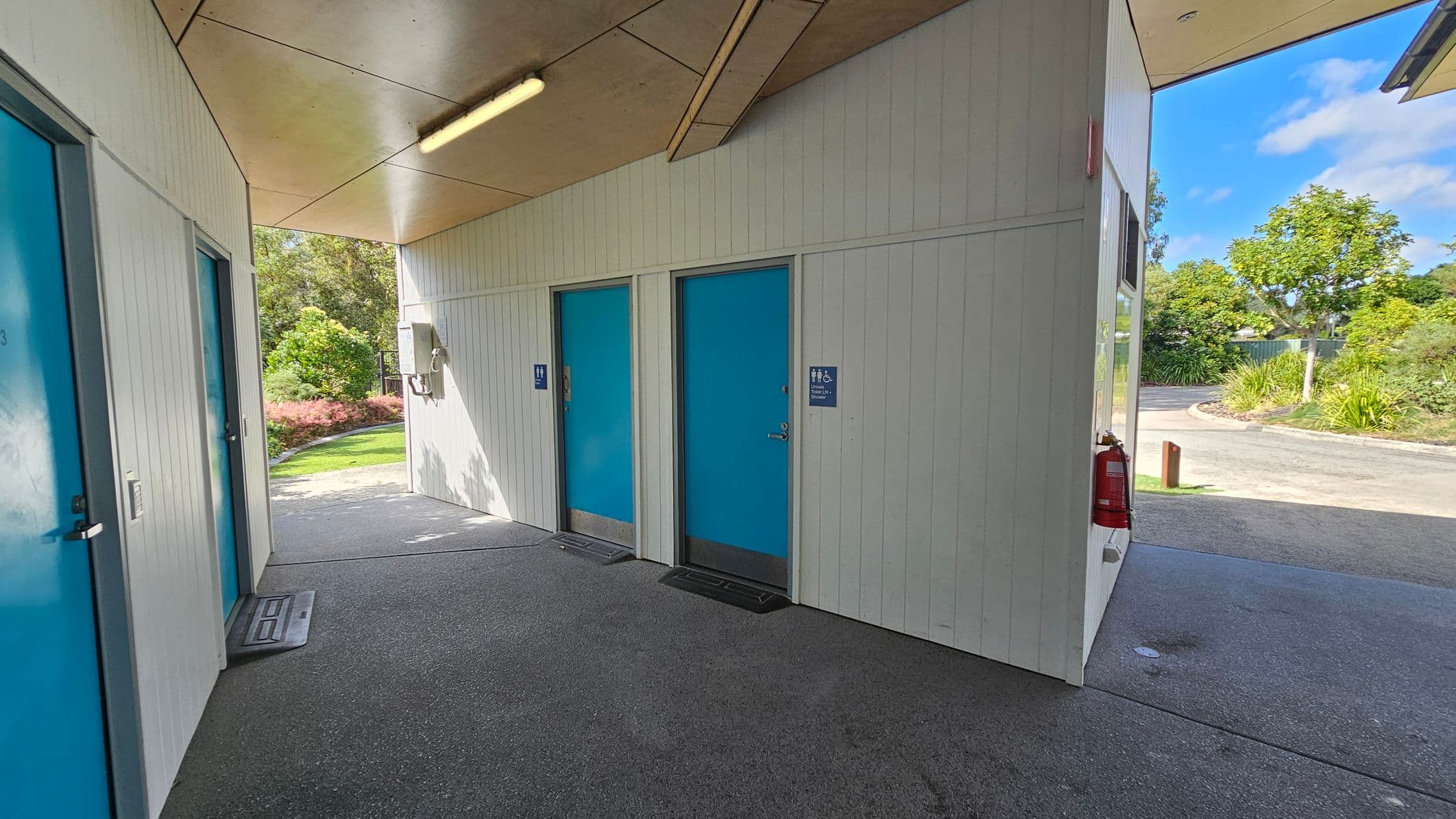 The space features a grey concrete floor that extends throughout the corridor. On the left, there are two doors, one marked with a blue sign indicating male facilities, while the other has a simple blue door. A light fixture mounted on the wooden panel ceiling illuminates the area evenly. To the right, the wall continues, and a fire extinguisher is visible near the corner. Beyond the exit, greenery provides a backdrop, with shrubs and a pathway visible through an opening leading outside.