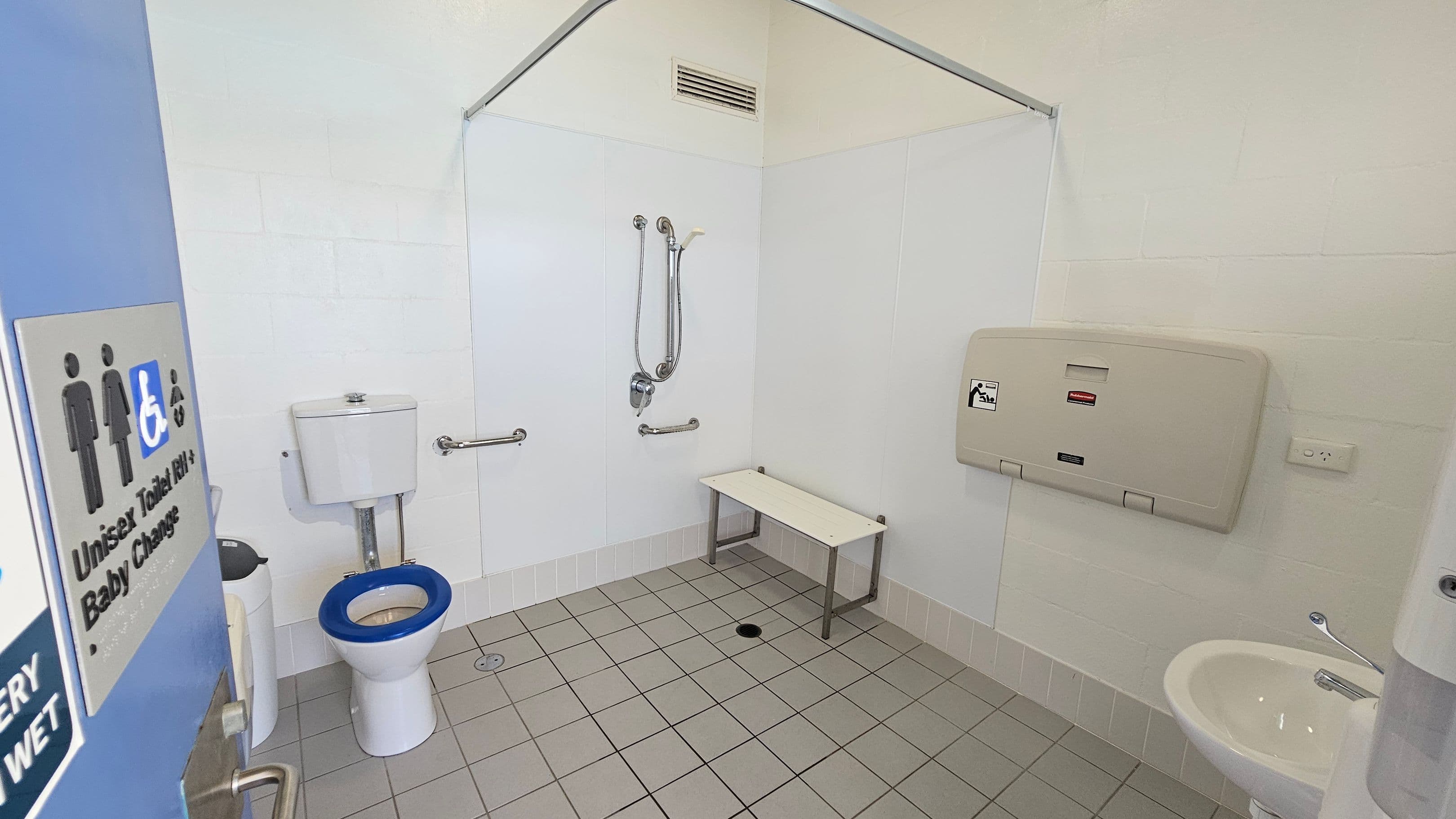 Accessible bathroom with a blue door on the left, featuring a sign indicating unisex and baby change facilities. Inside, a toilet with a blue seat is on the left, equipped with grab bars. A shower area with a handheld showerhead and additional grab bars is in the centre. A white bench is next to the shower. On the right, a baby changing station is mounted on the wall above a small sink. The floor is tiled in grey, and the walls are light-coloured.