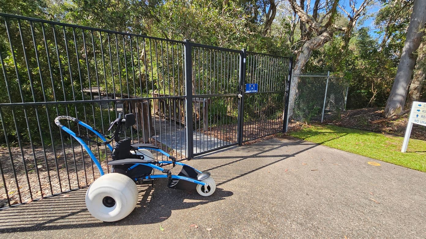 A smooth dark grey pathway leads up to a double black gate, which is partially open, allowing access to a wooded area beyond. On the left, a wheeled device sits on the path, featuring a white wheel and a blue frame, positioned close to the gate. To the right, a chain-link fence borders the pathway, and a sign indicating directions stands nearby. Lush green foliage surrounds the gate, providing a natural backdrop, while a variety of trees are visible beyond the fence.