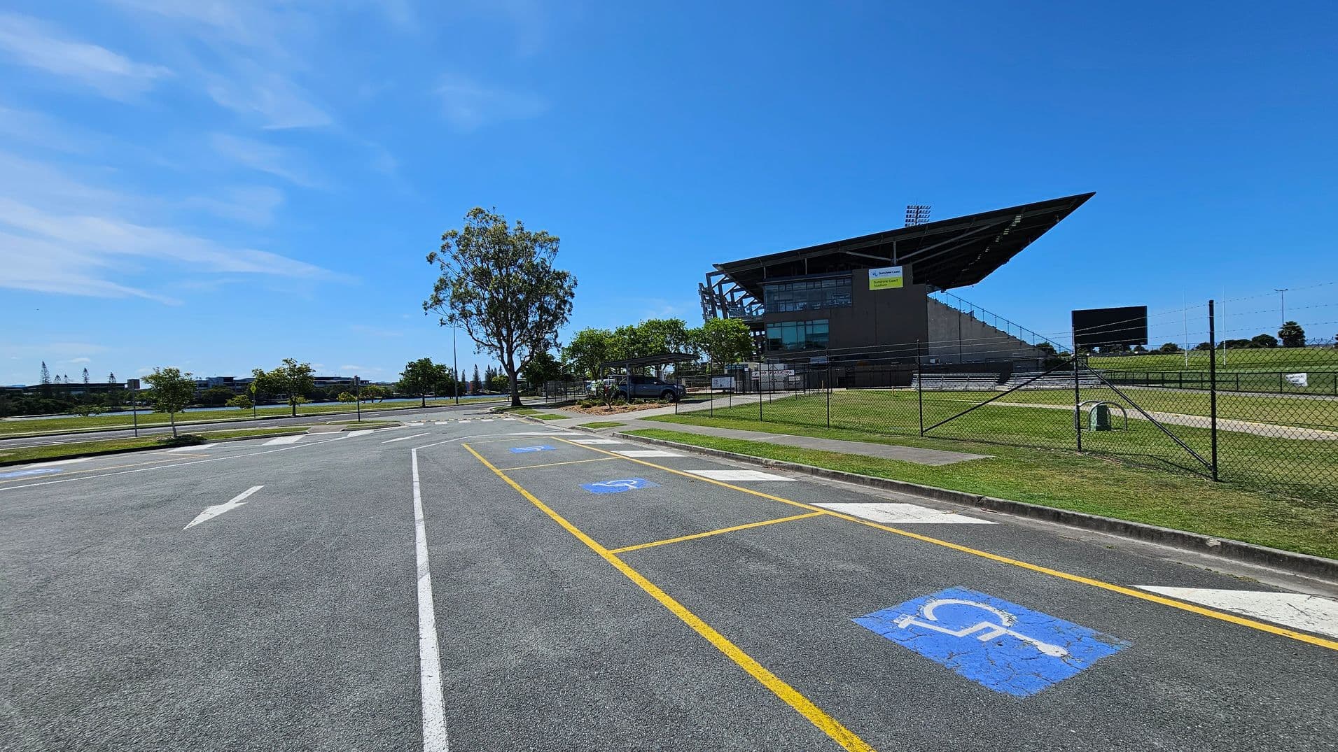 A large sports stadium is on the right, with a modern design and a prominent overhanging roof. In the foreground, there is a car park with marked disabled parking spaces, indicated by blue symbols on the asphalt. A chain-link fence runs along the perimeter of the stadium grounds. Trees and grass are visible around the area, with a few small trees lining the road on the left. The road has directional arrows painted on it.