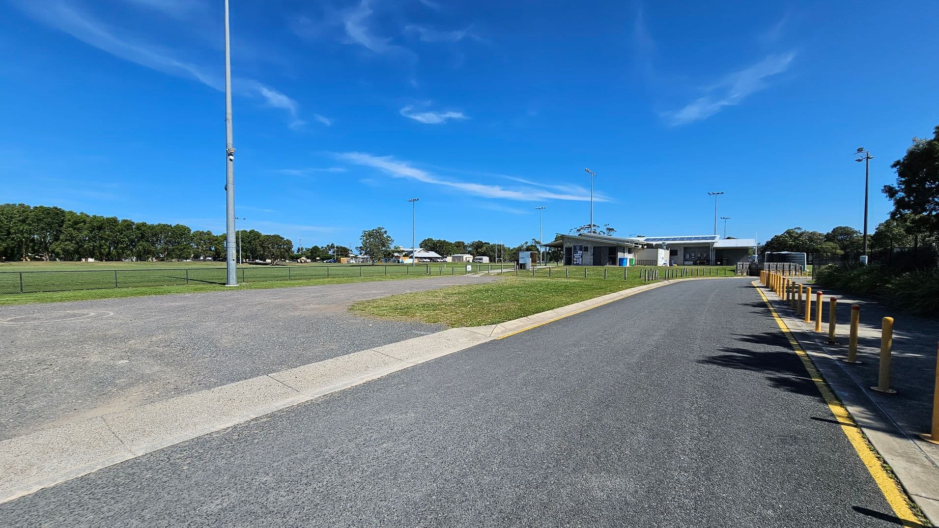 A paved road on the right with yellow bollards leads to a building in the distance. On the left, there's a grassy field bordered by a low fence and a tall light pole. Trees line the background, and the sky is clear.