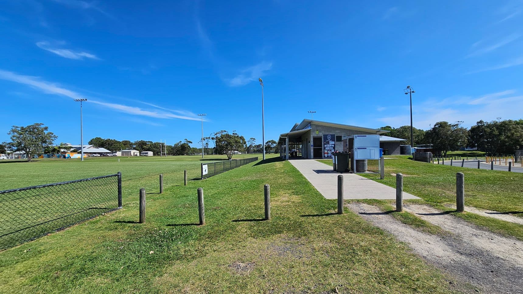A grassy sports field with a chain-link fence on the left. In the centre, a path leads to a building with a sloped roof and several signs on its walls. Tall floodlights are visible around the field. Wooden posts line the path, and there are trees and additional buildings in the background. The sky is clear and blue.