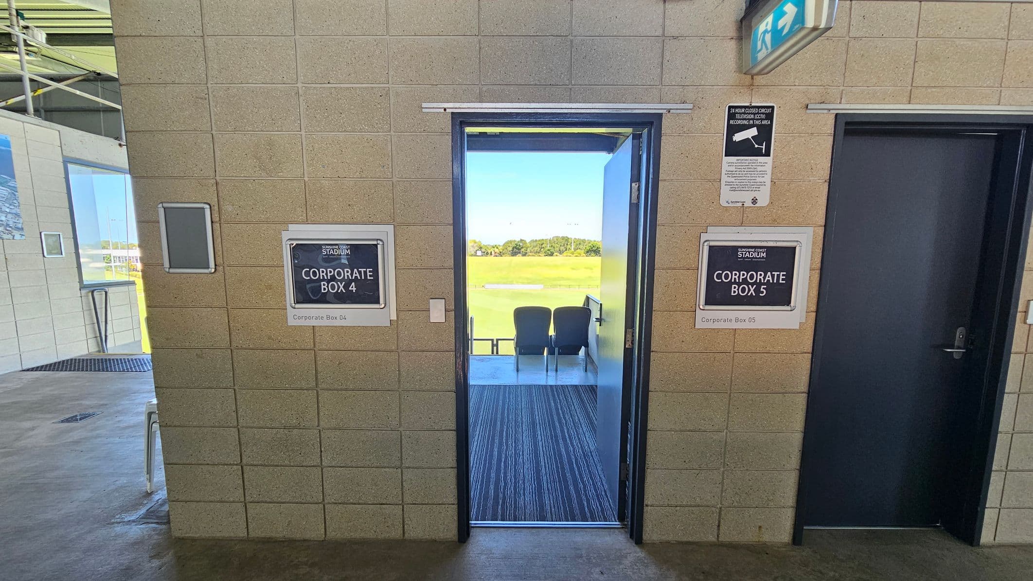A corridor with beige tiled walls. In the centre, an open door leads to Corporate Box 4, revealing two chairs facing a sports field. To the right, a closed door marked Corporate Box 5. Above, a CCTV sign and an emergency exit sign. On the left, part of another room with a window and a chair visible. The floor is concrete, and the ceiling has visible metal beams.