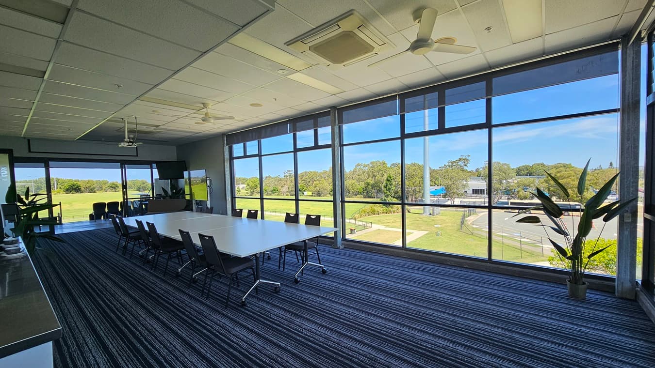 A conference room with large floor-to-ceiling windows on the right, offering a view of a green landscape and trees. In the centre, there is a long white table surrounded by black chairs on a striped carpet. On the left, there is a counter with cups and a plant. The ceiling has a projector and ceiling fans. A potted plant is near the windows.