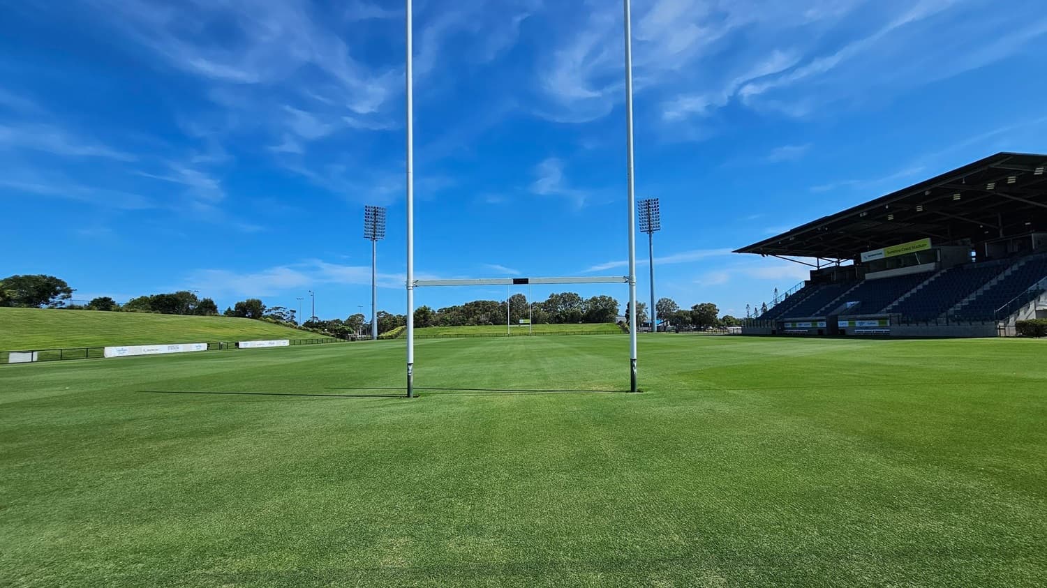 A rugby field with bright green grass. In the centre, there are rugby goalposts. On the right, a large covered grandstand with tiered seating. On the left, a grassy embankment with advertising boards. Tall floodlights are positioned on both sides of the field. Trees and shrubs are visible in the background.