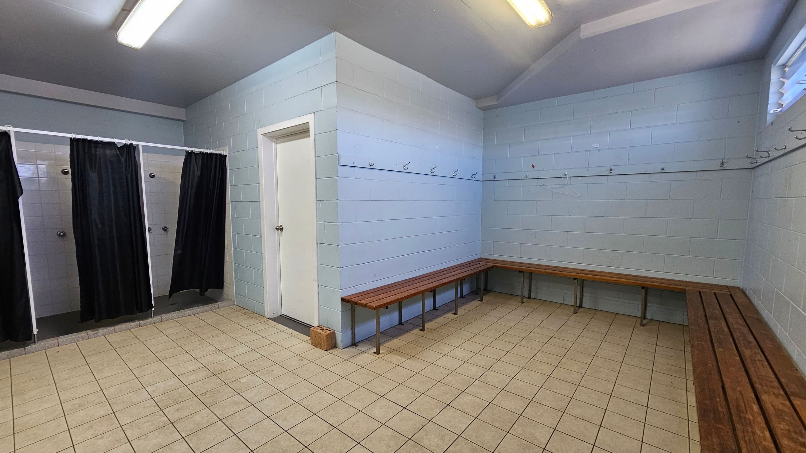 A changing room with light blue and white tiled walls. On the left, there are three shower stalls with black curtains. In the centre, a white door is visible. On the right, wooden benches line the walls, with hooks above them for hanging items. The floor is covered with beige tiles, and the ceiling has fluorescent lights. A small window is on the upper right wall.