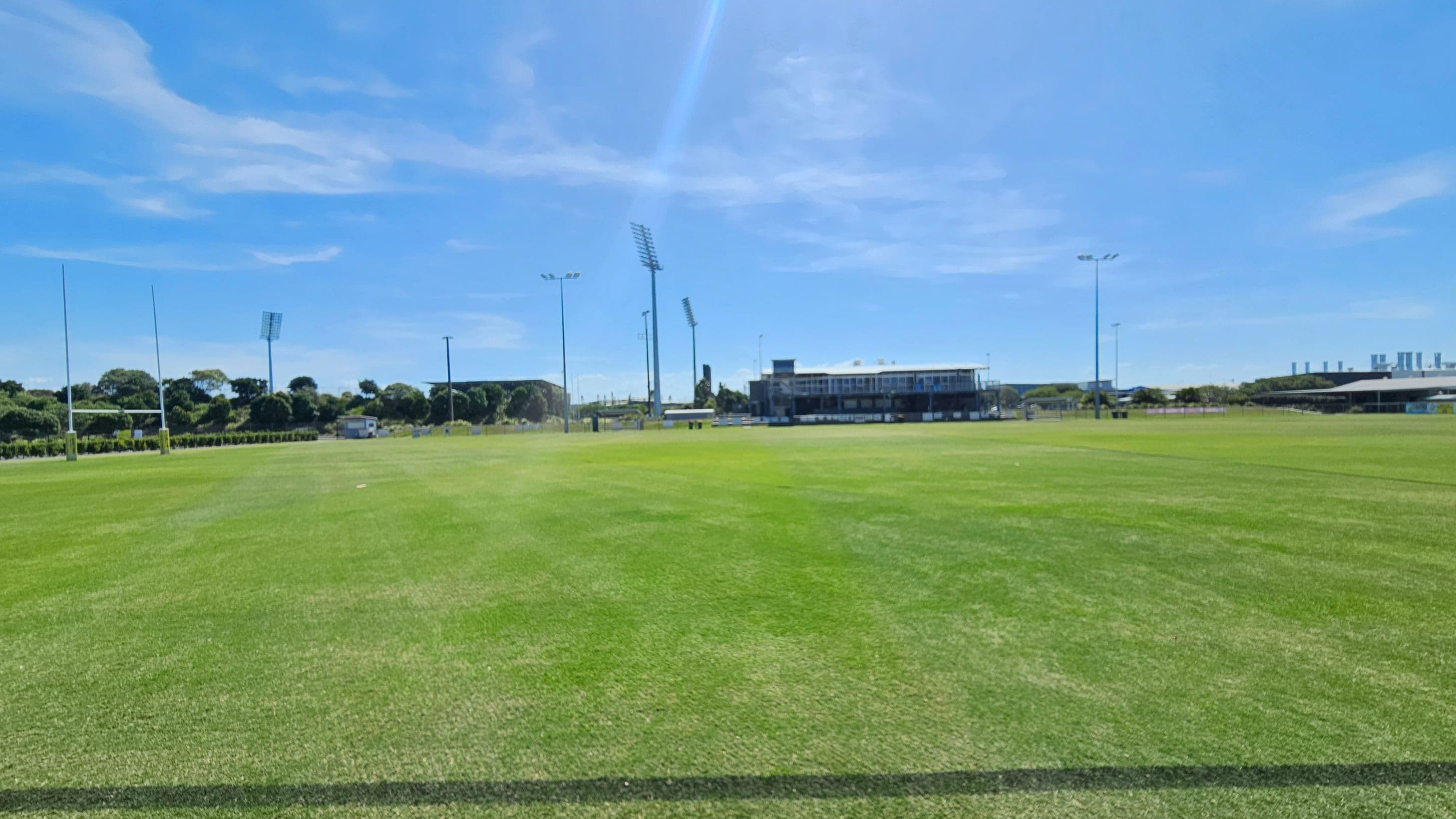 A large, well-maintained grassy sports field with rugby posts on the left. In the background, there are several tall floodlights and a building with a viewing area. Trees and some industrial structures are visible in the distance. The field is expansive, with clear lines marking the playing area.