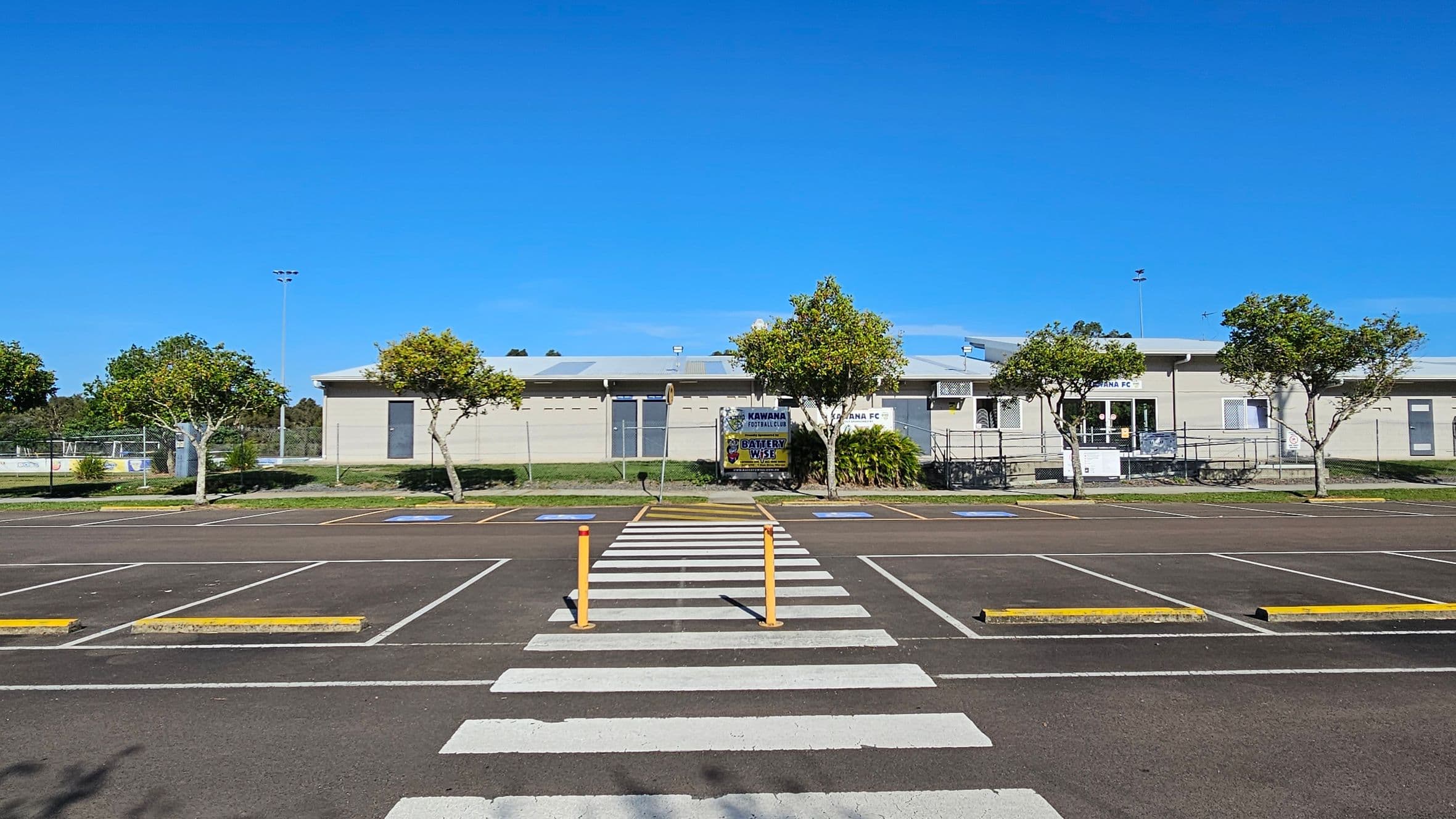 A parking area with marked spaces and a zebra crossing in the foreground. Two yellow bollards are positioned at the crossing. In the background, a single-storey building with a light-coloured facade is visible, flanked by several small trees. Signs on the building read "Kawana Football" and "Kawana FC". The sky is clear and blue.