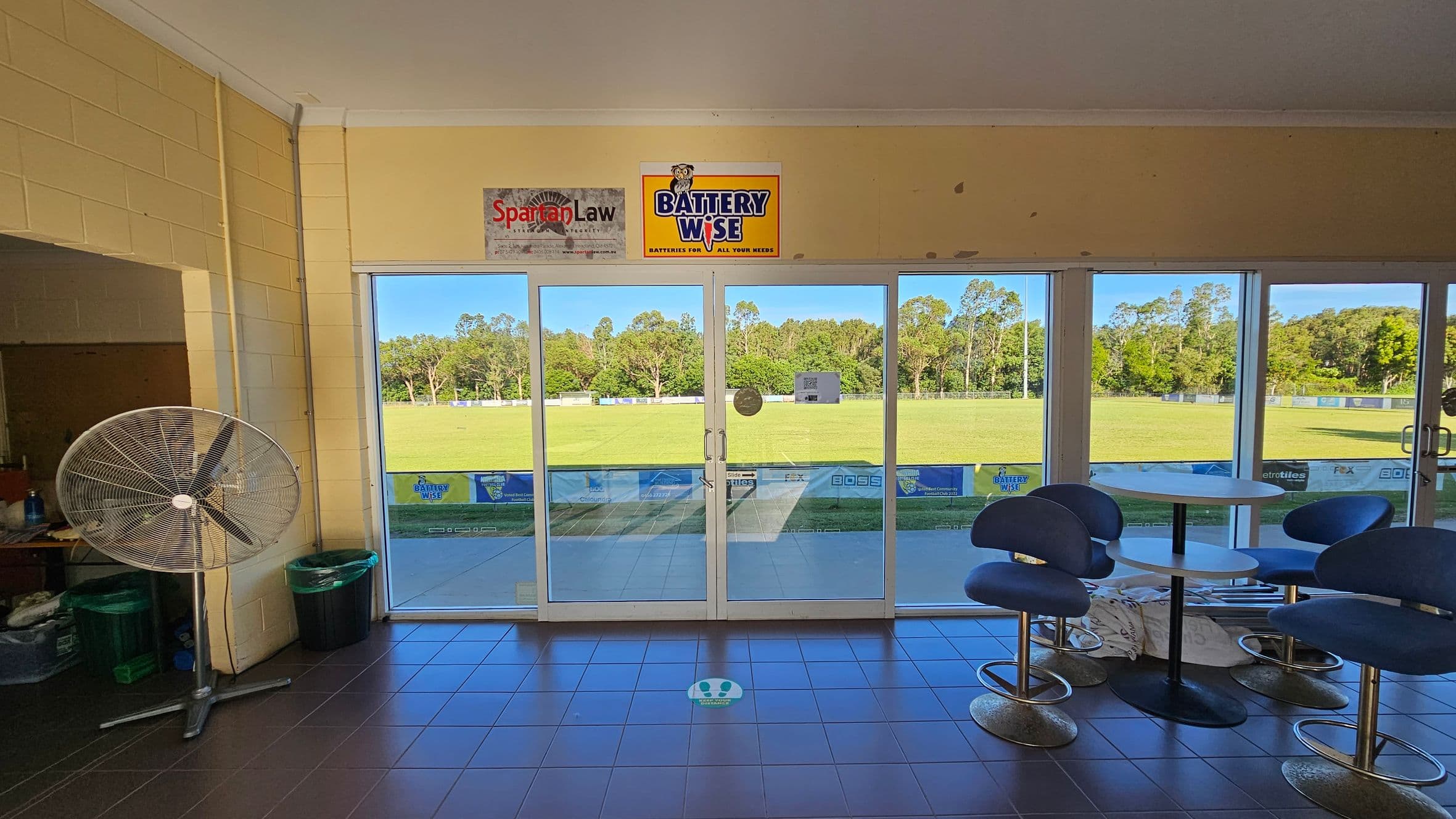 A room with large glass doors in the centre, leading to a grassy field outside. On the left, there's a large standing fan and a couple of bins. On the right, there are several blue chairs around a round table. The floor is tiled in dark grey, and there are two signs above the doors, one for "Spartan Law" and another for "Battery Wise". Trees are visible in the background through the glass doors.