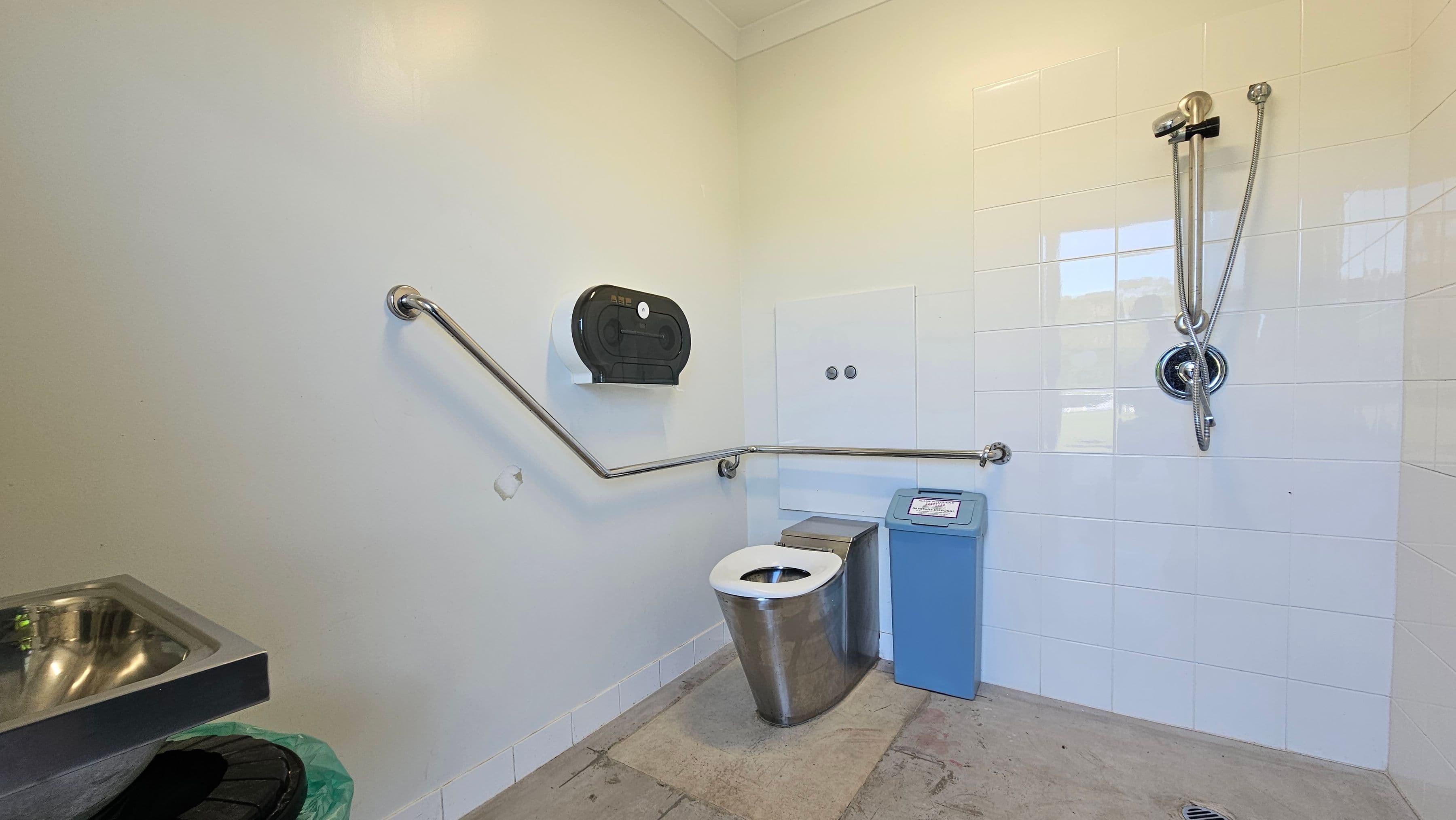 A public restroom with a stainless steel toilet in the centre, equipped with a blue sanitary bin to the right. The walls are partially tiled in white, with a shower fixture on the right wall. A horizontal grab rail runs along the walls, and a black toilet paper dispenser is mounted above the toilet. On the left, a stainless steel sink is visible, with a green bin beneath it. The floor is concrete, and the ceiling is plain.