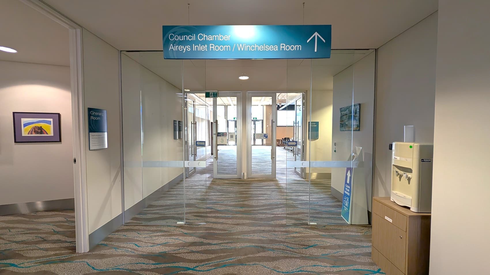 A hallway with a patterned carpet leads to glass doors. Above the doors, a sign indicates directions to the Council Chamber, Aireys Inlet Room, and Winchelsea Room. On the left, there's a doorway to the Gherang Room with a colourful painting on the wall. On the right, a water dispenser sits on a wooden cabinet. Beyond the glass doors, a larger room is visible with more patterned carpet and some furniture.