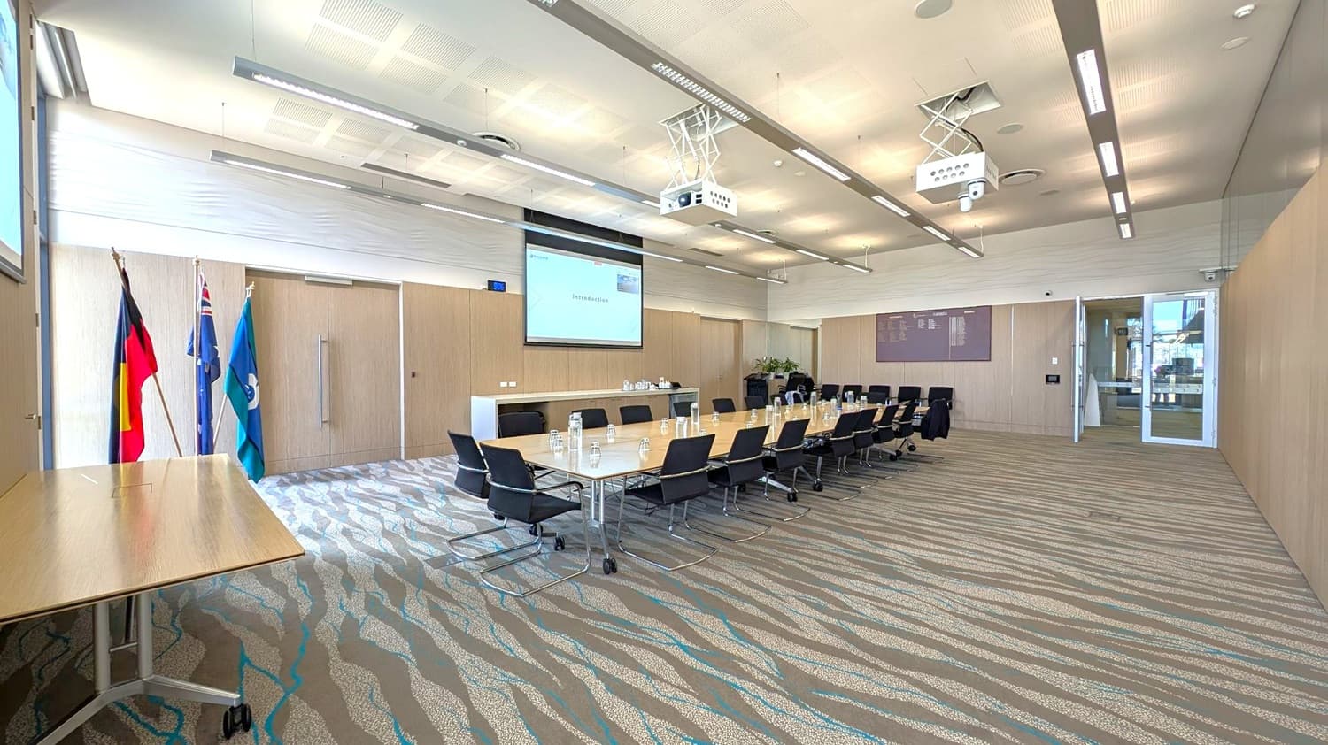 A conference room with a long rectangular table in the centre, surrounded by black chairs. On the left, there are three flags next to a small table. The right side features a glass door leading outside. The ceiling has recessed lighting and two projectors. The floor is carpeted with a wavy pattern in green, brown, and blue. A large screen at the front displays a presentation titled "Introduction." A sideboard with water bottles and glasses is against the wall.