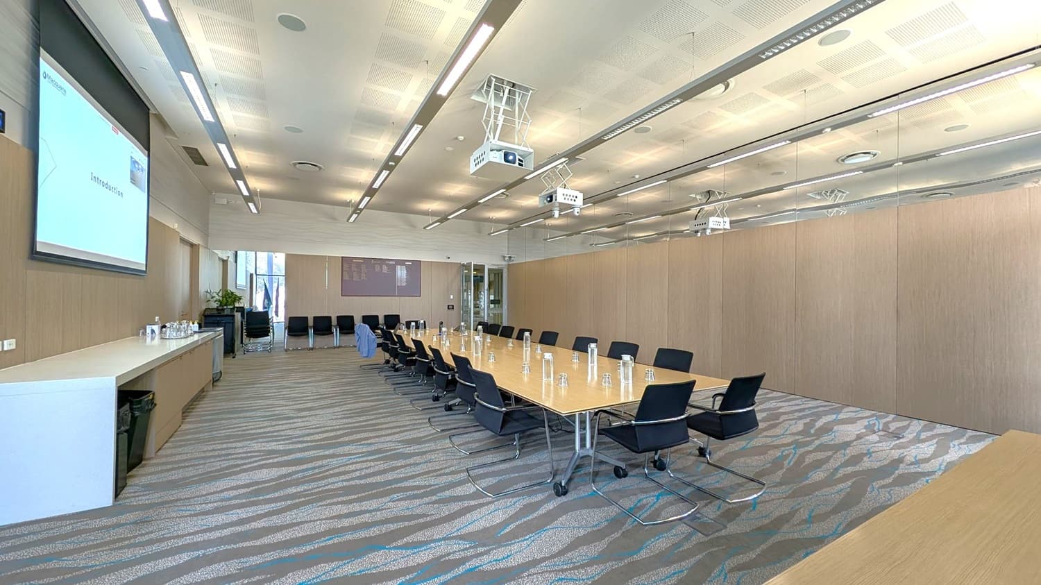 A modern conference room with a long rectangular table in the centre, surrounded by black chairs. On the left, a large screen displays a presentation titled "Introduction." Below the screen, a counter holds water bottles and glasses. The right wall is lined with wooden panels. The ceiling features recessed lighting and two projectors. The floor has a patterned carpet in grey and blue tones. At the back, a glass door and additional chairs are visible.