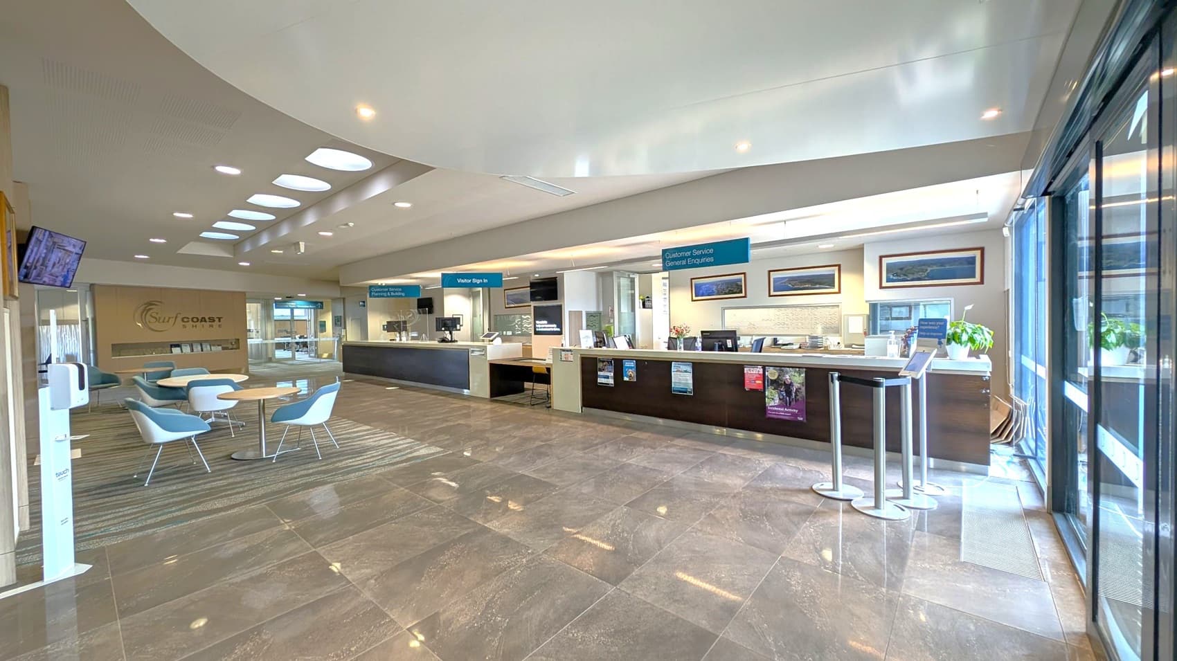Automatic glass doors open to reveal an interior space with a tiled floor. Inside, there are chairs and a table in the centre, with a TV mounted on the left wall. A reception desk is on the right, where a person is standing. The ceiling has circular lights, and there are large windows on the right side, allowing natural light to enter. A sign on the window mentions COVID-19 testing.