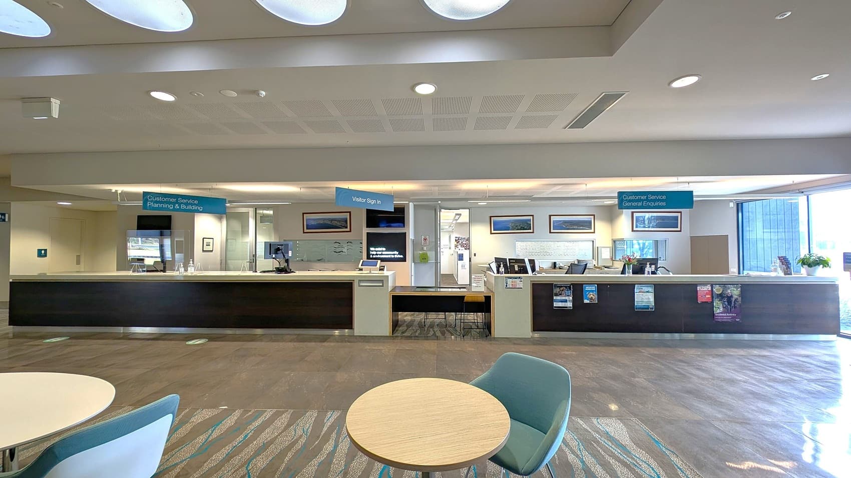 A spacious reception area with a long counter in the centre, divided into sections for "Customer Service Planning & Building," "Visitor Sign In," and "Customer Service General Enquiries." Behind the counter are computers and framed pictures on the wall. The floor is a mix of wood and carpet, and the ceiling has circular light fixtures. In the foreground, there is a small round table with a green chair. Large windows on the right let in natural light.