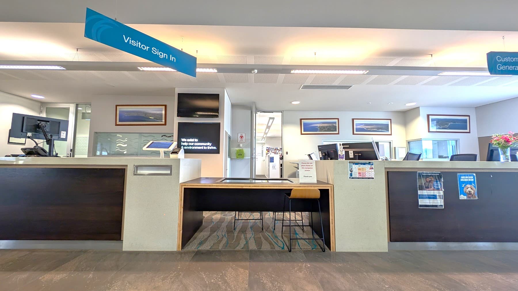 A reception area with a counter in the centre, featuring a digital sign and a tablet. Above, a sign reads "Visitor Sign In". On the left, a computer monitor is visible. The right side has another counter with a computer and a vase of flowers. The background shows framed pictures on the wall and a doorway leading to an office space. The floor is carpeted with a patterned design, and the ceiling has recessed lighting.