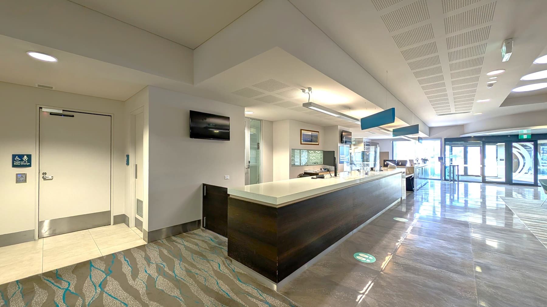A modern reception area with a long, dark counter on the right. Behind the counter are computer monitors and a glass partition. On the left, there's a door with a sign indicating accessibility and baby changing facilities. Above, the ceiling has recessed lighting and a suspended light fixture. The floor is a mix of patterned carpet and shiny tiles. In the background, large glass doors lead outside, allowing natural light to flood the space.