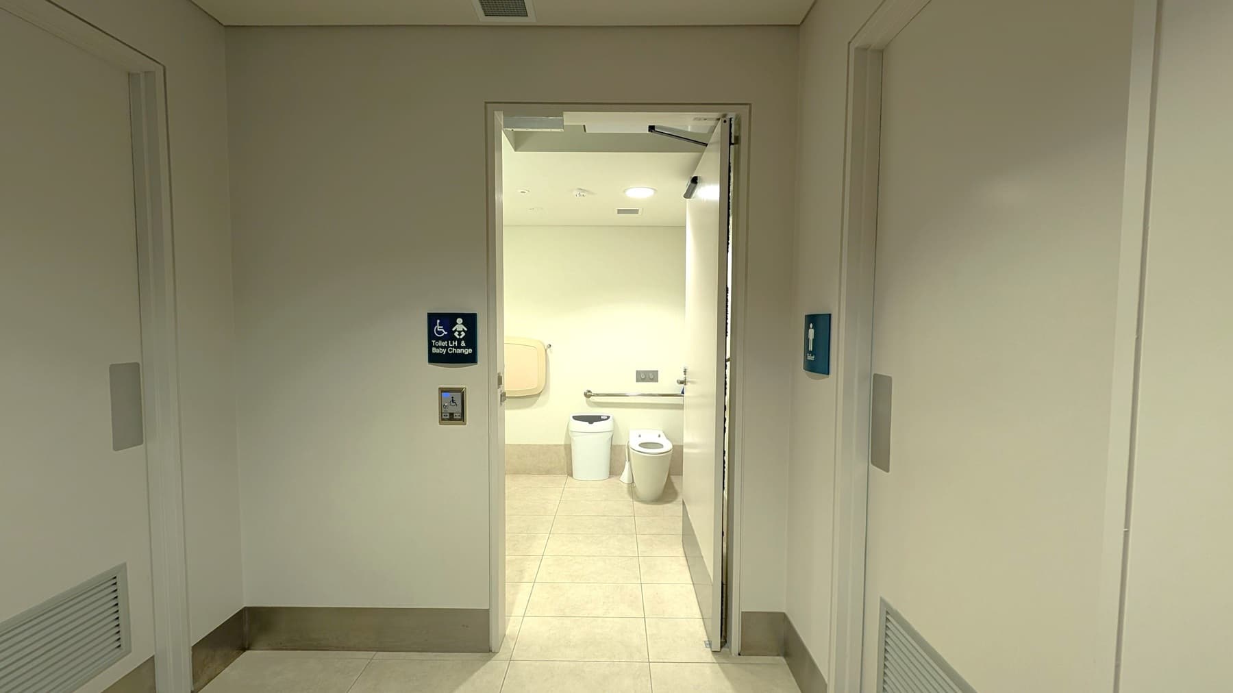 A hallway with beige walls and a tiled floor leads to an open door revealing a bathroom. The bathroom has a beige tiled floor and a white toilet with a bin next to it. A baby changing station is mounted on the left wall. Signs on the door indicate accessibility features, including a baby change facility. The ceiling is plain with recessed lighting.