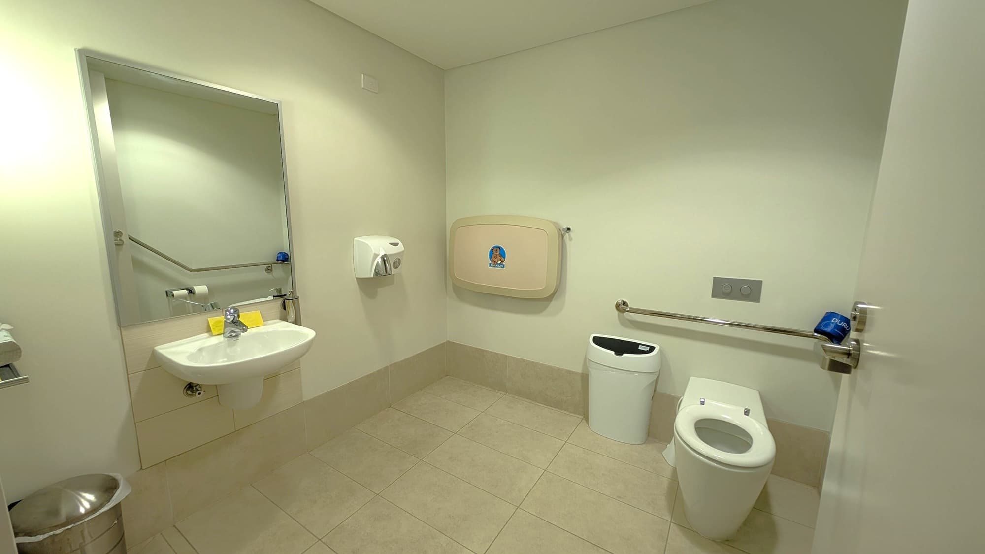 A public restroom with a white toilet on the right, equipped with a grab bar and a blue roll of toilet paper. A white bin is next to the toilet. On the left, a wall-mounted sink with a mirror above it, a soap dispenser, and a hand dryer. A baby changing station is mounted on the wall in the centre. The floor is tiled in light brown, and the walls are a light grey.