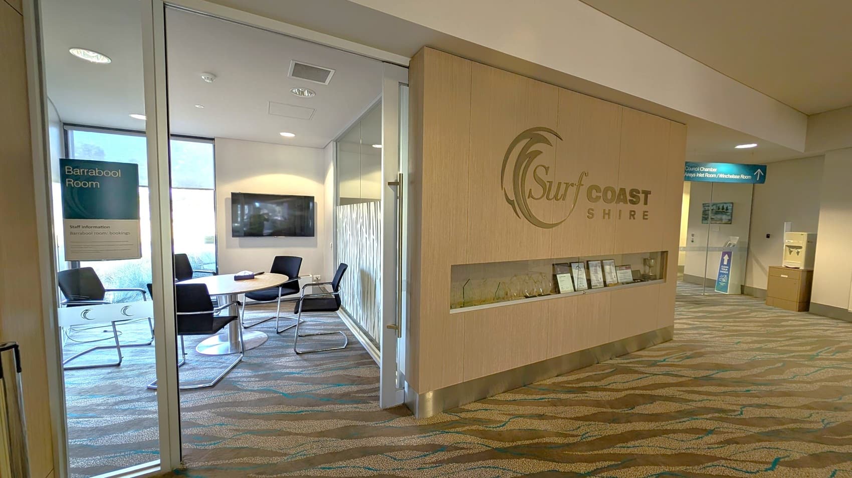 A modern office interior with a glass-walled meeting room on the left, labelled "Barrabool Room." Inside, there is a round table surrounded by black chairs and a wall-mounted TV. On the right, a wooden wall features the "Surf Coast Shire" logo and a display shelf with awards. The carpet has a wavy pattern, and there are directional signs in the hallway leading to other rooms. The ceiling is fitted with recessed lighting.