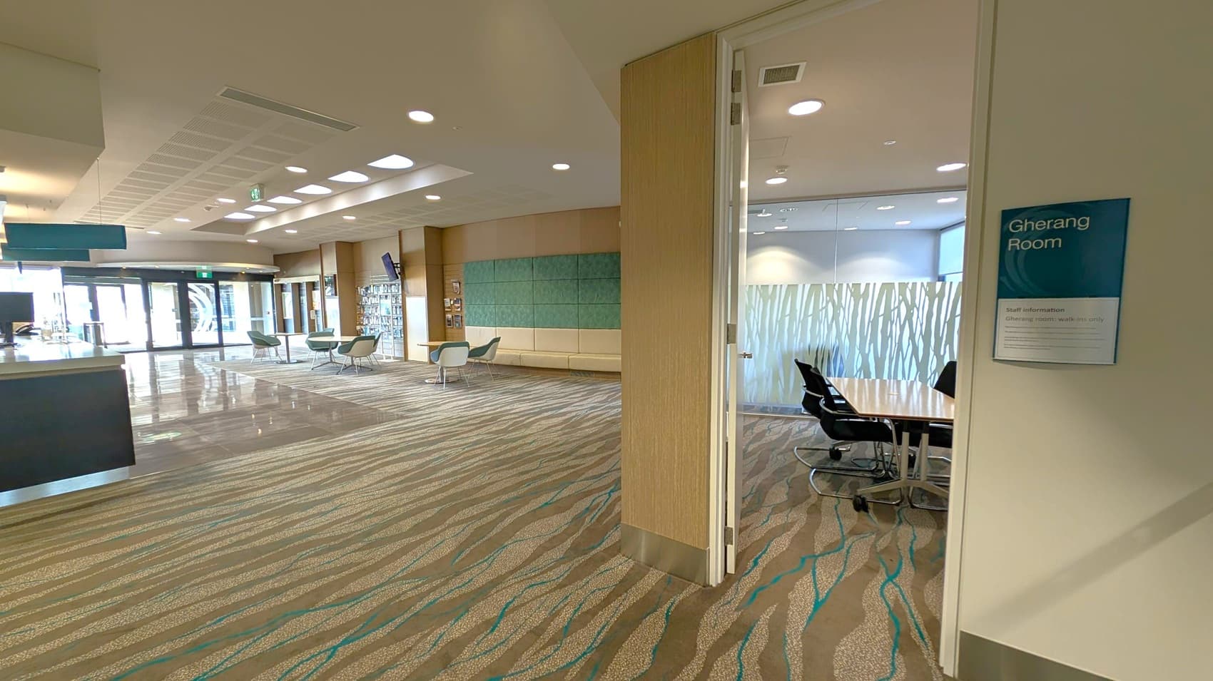A modern interior space with a patterned carpet in shades of brown and green. On the right, there's an open door leading to the "Gherang Room," which has a glass wall with a frosted design and a table with black chairs. In the background, the main area features seating with green chairs and a wall with blue and green panels. The ceiling has recessed lighting and skylights, and there are large glass doors at the far end.