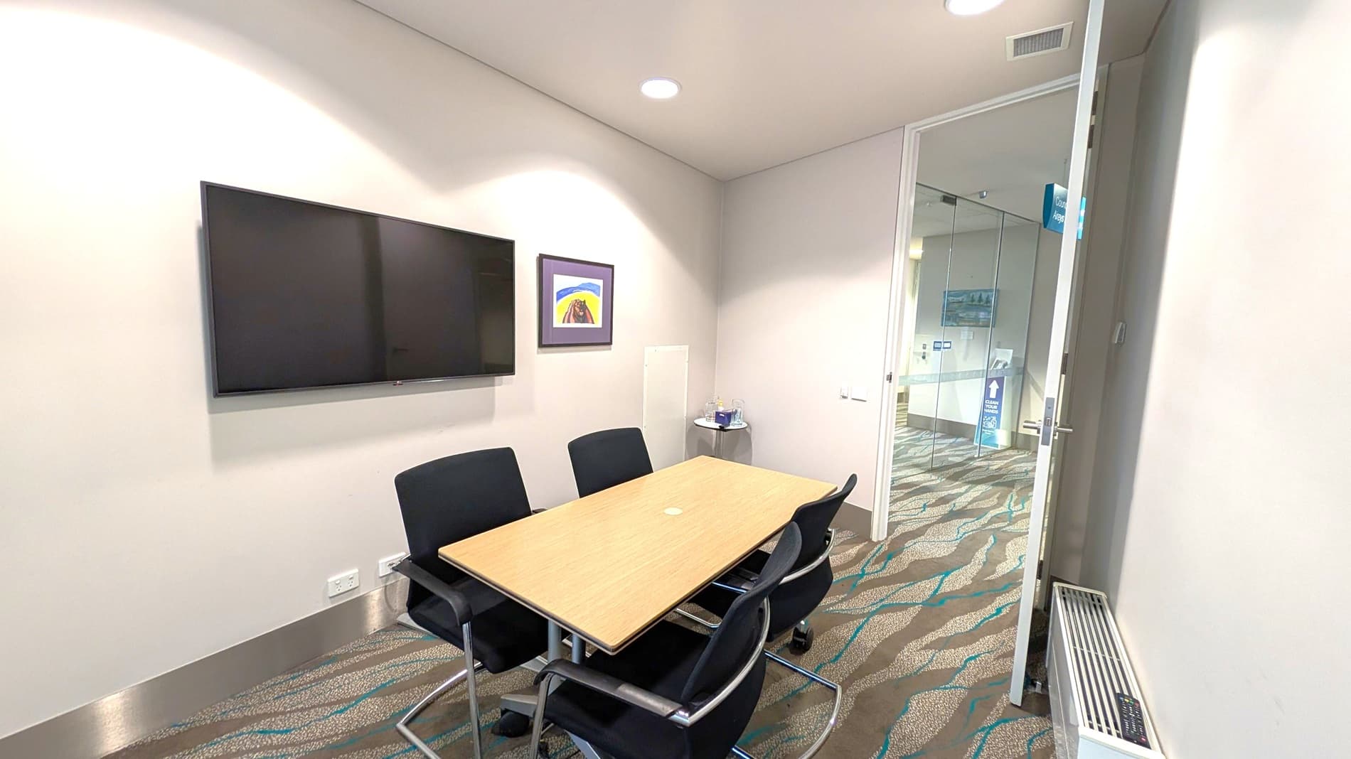 A small meeting room with a rectangular wooden table surrounded by four black chairs. On the left wall, a large flat-screen TV is mounted, with a framed picture below it. In the corner, a small round table holds a water bottle and glasses. The floor has a patterned carpet in shades of brown and blue. The ceiling is white with recessed lighting. A glass door on the right leads to a hallway with a sign visible through the glass.