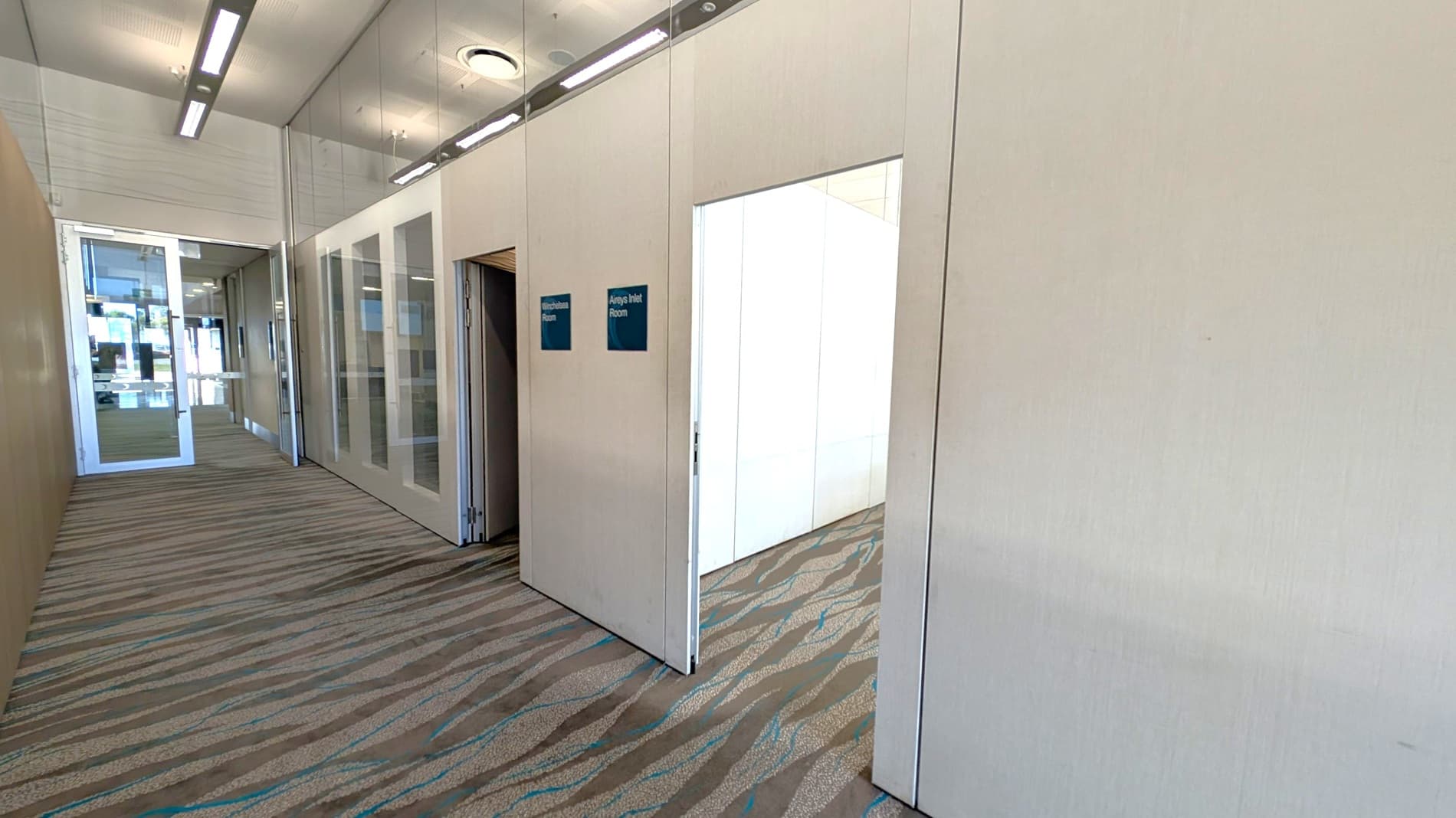 A hallway with a patterned carpet floor and a ceiling with recessed lighting. On the left, there are glass doors leading to another area. On the right, there are two open doors with signs above them reading "Birchbark Room" and "Aireys Inlet Room." The walls are light-coloured, and the rooms beyond the doors have similar patterned carpets.