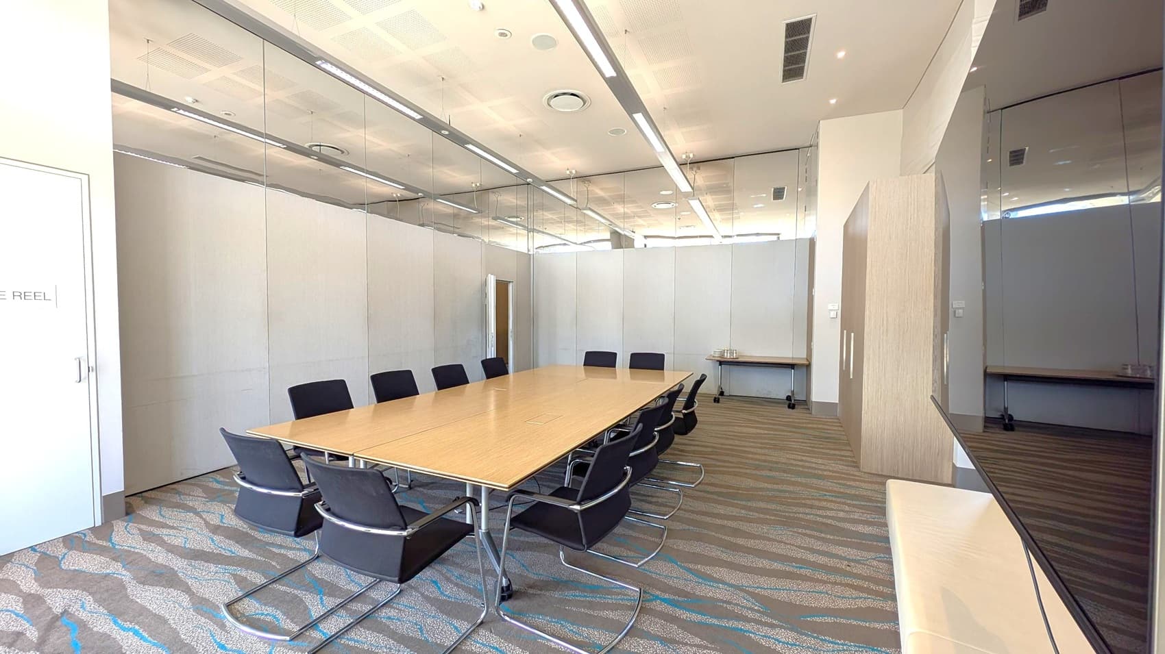 A conference room with a long wooden table in the centre, surrounded by black chairs. The left side has a door with a sign, and the right side features a mirrored wall. The back wall is plain with a small table holding a few items. The floor is carpeted with a wavy pattern, and the ceiling has recessed lighting and air vents.