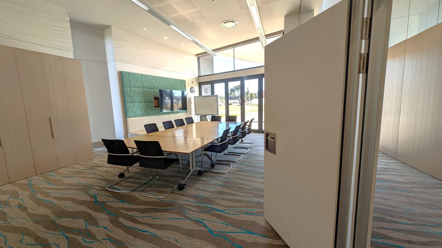 A modern conference room with a large rectangular table surrounded by black chairs. On the left, there are wooden cabinets. The back wall features a mounted flat-screen TV and a clock. A whiteboard stands near the glass doors on the right, which offer a view of the outdoors. The floor is carpeted with a wavy pattern, and the ceiling has recessed lighting and air vents.