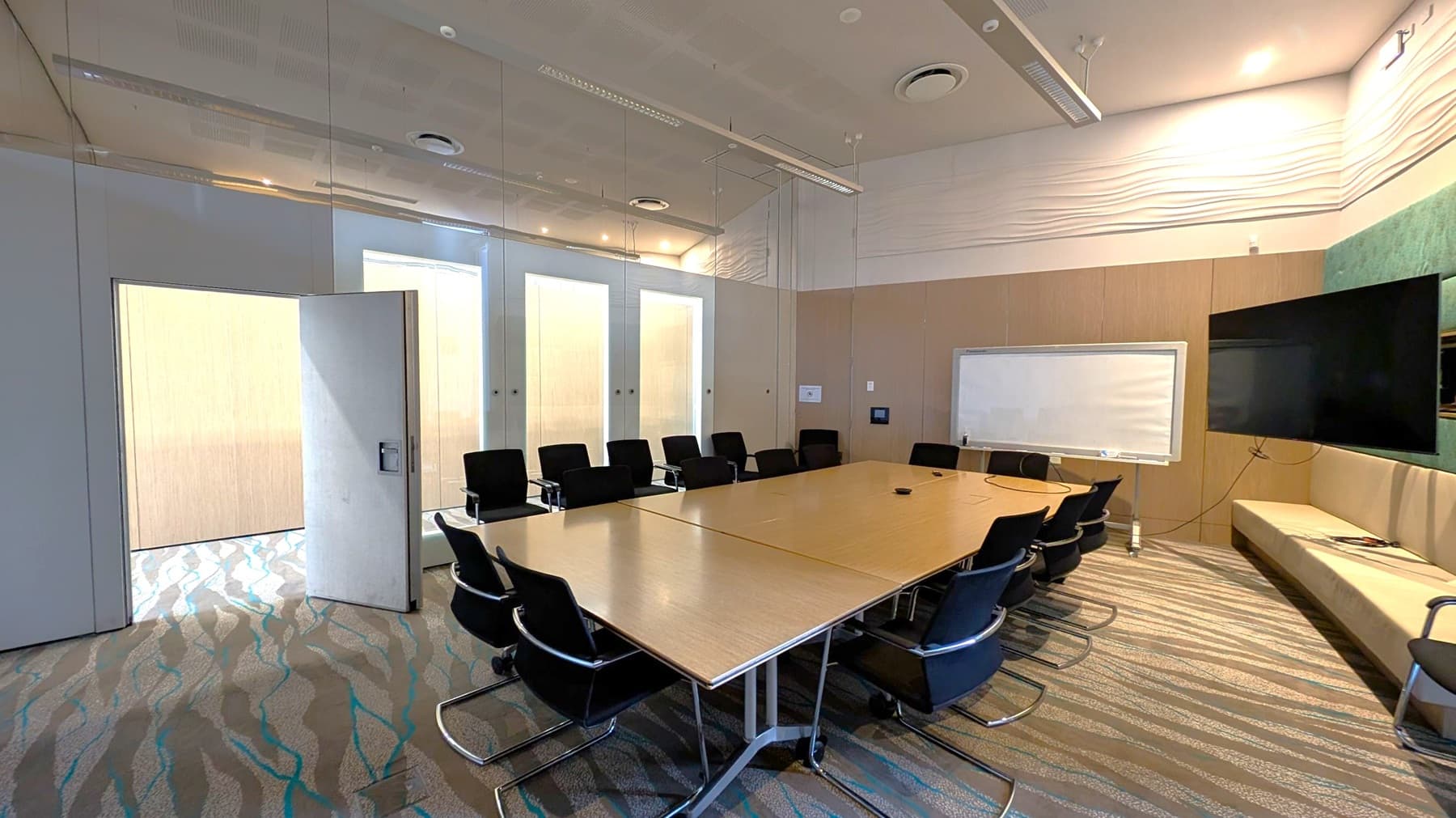 A modern conference room with a large rectangular table surrounded by black chairs. On the right, there's a wall-mounted flat-screen TV and a whiteboard. The walls are light-coloured with a textured design near the ceiling. The floor is carpeted with a wavy pattern. On the left, a door is slightly open, revealing a bright area outside. The ceiling has recessed lighting and air vents.