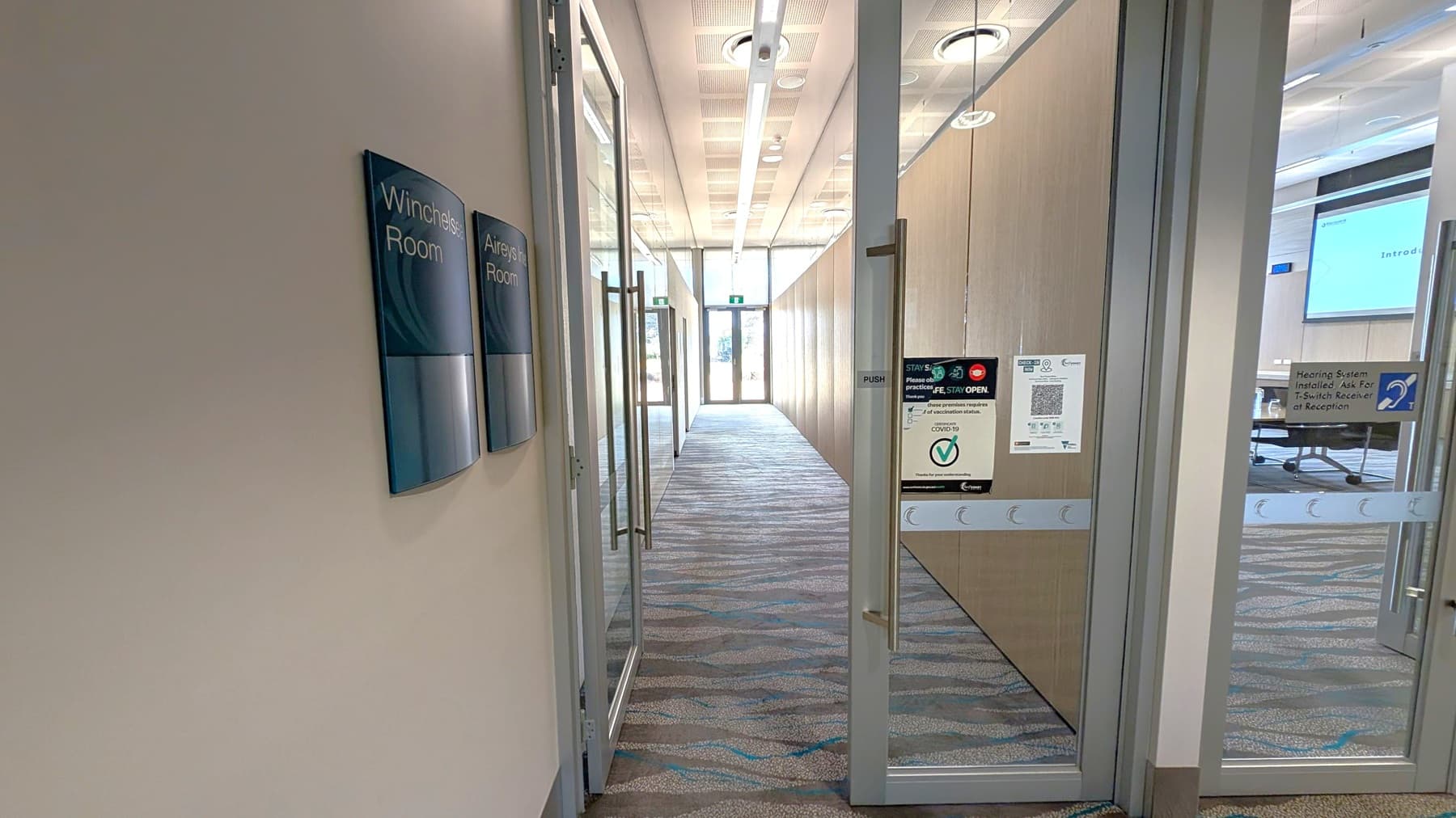 A corridor with a patterned carpet floor and a ceiling with circular lights. On the left, there are signs for "Winchelsea Room" and "Aireys Inlet Room" on the wall. On the right, glass doors with various notices, including COVID-19 guidelines and a hearing system notice. Through the glass, a room with a screen and tables is visible. At the end of the corridor, there is a glass door leading outside, allowing natural light to enter.