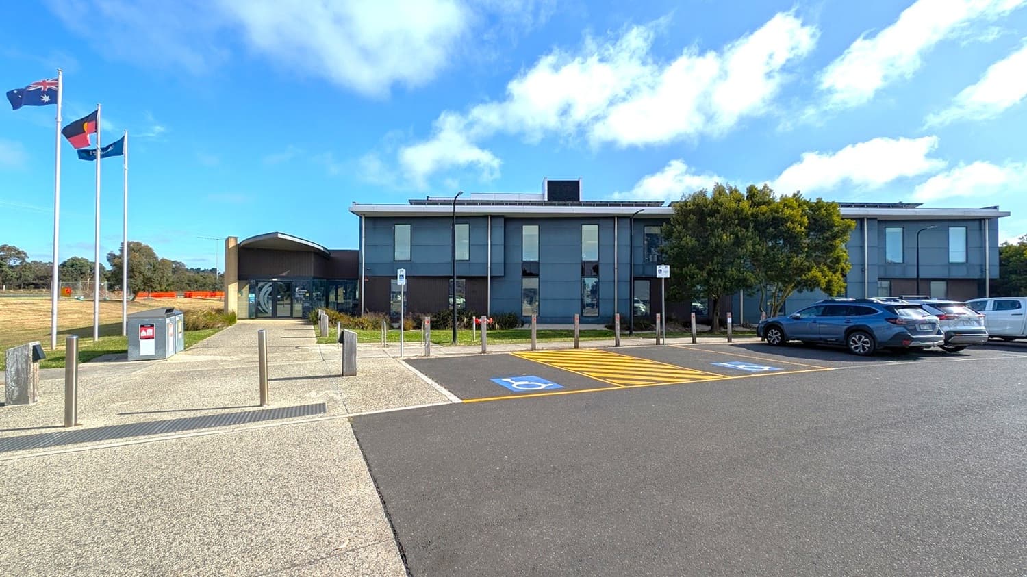 A modern building with large windows is on the right, surrounded by a car park with several vehicles. In the foreground, there are marked disabled parking spaces with blue and yellow lines. Three flagpoles with flags are near the building. On the left, there's a grassy field with scattered trees and a few orange barriers. The sky is clear with some clouds.