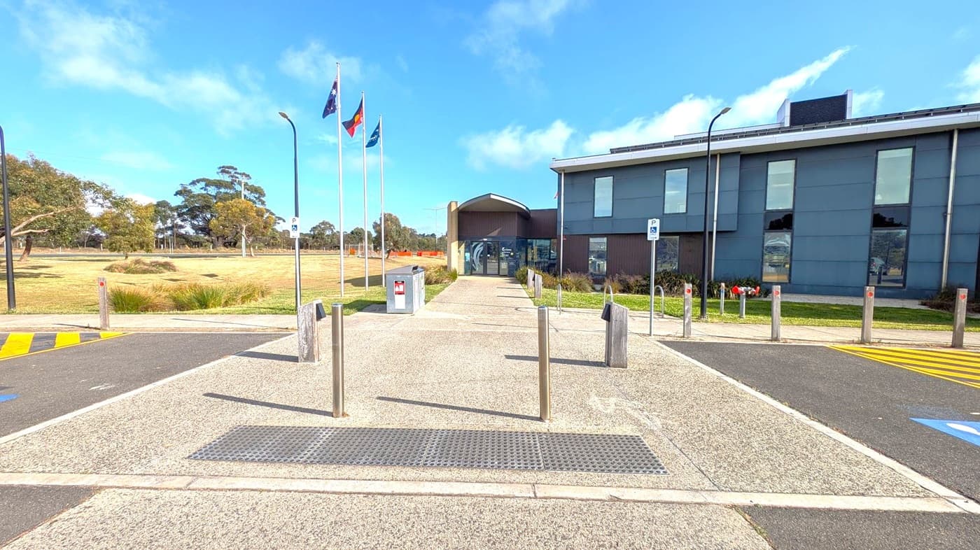 A paved pathway leads to a modern building with large windows on the right. Three flagpoles with flags are on the left, next to a grassy area with trees. The foreground features a tactile paving strip and several bollards. A parking sign is visible near the building, and the road has yellow markings.
