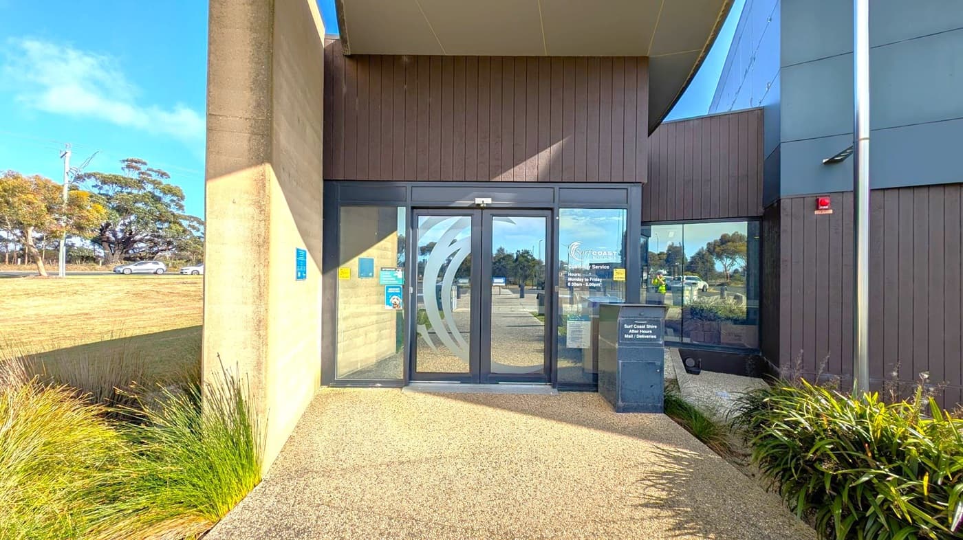 Entrance to a modern building with glass doors featuring a logo and business hours. The left side has a concrete wall with a blue sign and green plants at the base. The right side has dark panelling and a small window. A metal post is visible near the corner. In the background, a road with a silver car and trees can be seen. The ground is covered with a textured surface.