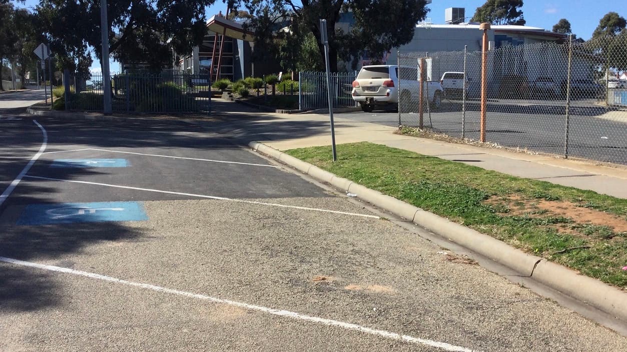 In close proximity to the centre’s main entrance is the carpark with accessible parking spaces. There is a chain link fence that serves as the carpark’s facade and a blue gate on its left where cars may enter to park. In front of the wired fence is a white pavement leading to several accessible car spaces. From this view, you will see the centre surrounded by trees.
