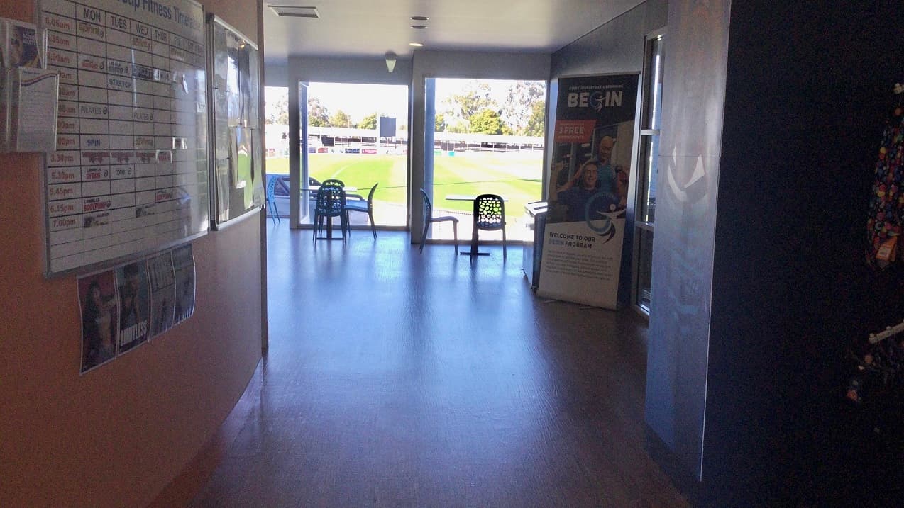 This is the extended section of the leisure centre entrance that highlights the orange wall that has the Fitness Timetable posted on the large white board. There are also flyer racks mounted on the wall where flyers are displayed. There are sets of tables and chairs placed near the glass walls where you will enjoy a pleasant green scenery. A standee of a human-sized poster is placed in the middle of this section where the word “BEGIN” is highlighted.