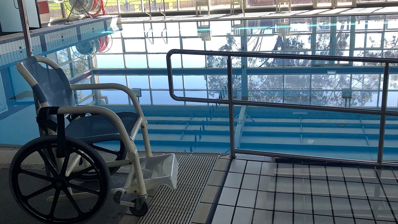 A closer look of the pool area where a blue and white aquatic wheelchair is placed on the side of the pool. The blue water shows the reflection of the white ceiling. The inner side of the pool’s coping has a text saying “Deep Water”. There are also silver pool stairs and an access ramp.