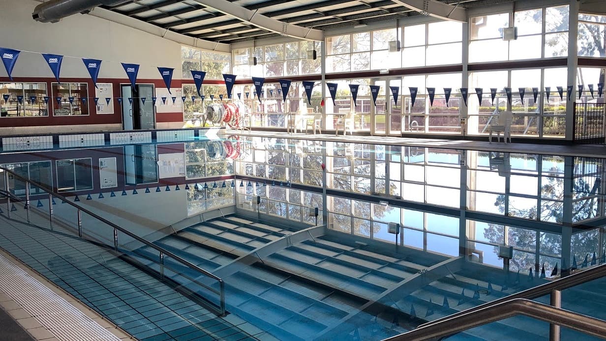 The view of the pool area where it’s showing the entire pool body. The blue water shows the reflection of the white ceiling and a long silver access ramp is at the side of the pool. There are long hanging banners made of white string and triangular blue pennants where “Zoggs“is printed. The glass wall of the pool area will let you see the bright and relaxing view from outside.
