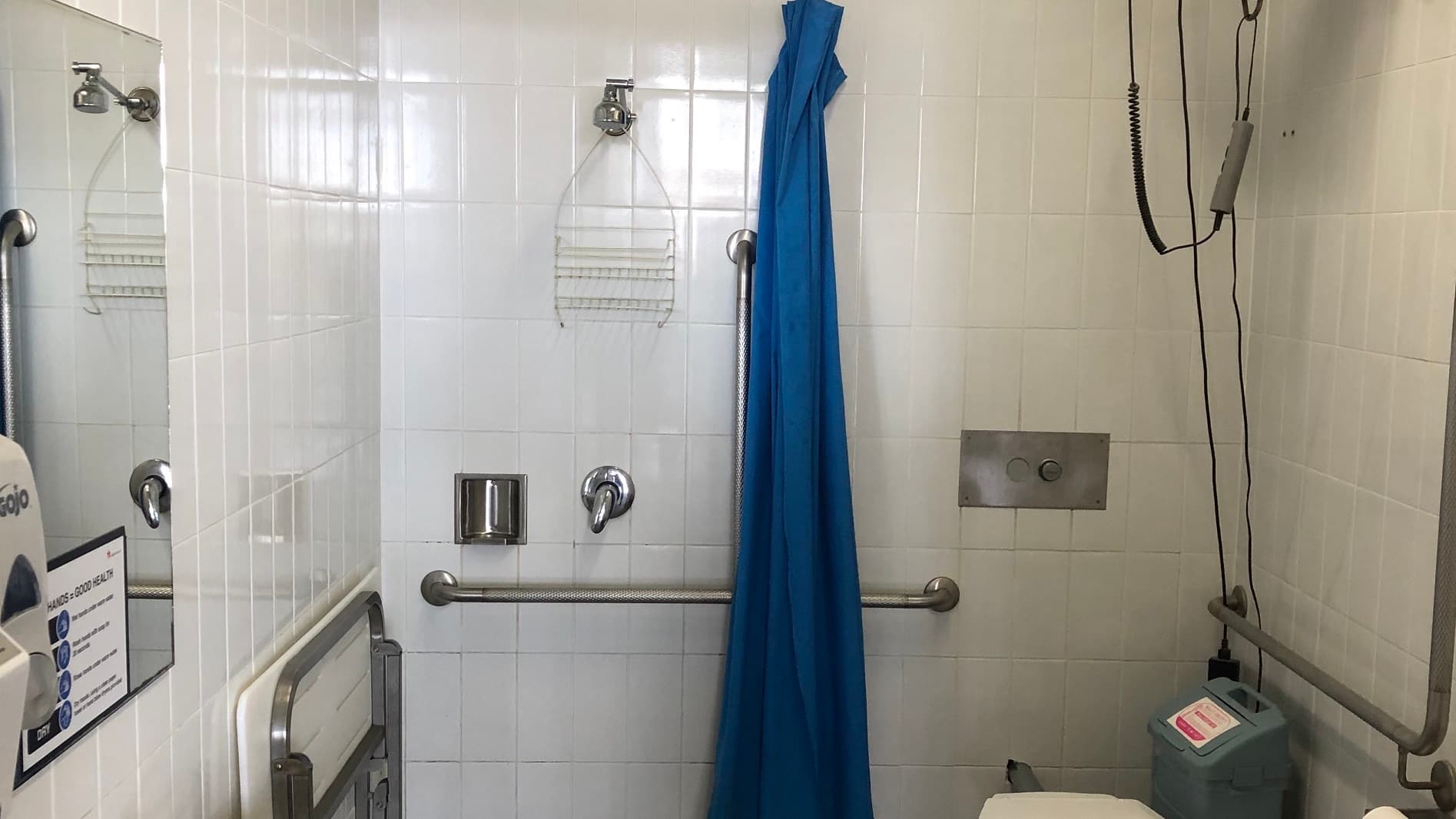 The leisure centre’s closer look of the white tiled restroom. It has silver grab handles at both sides, a silver built-in soap holder and a hanging accessory rack. A rectangular mirror is mounted on the left wall with a reminder posted on the bottom about the importance of washing hands. A blue folded curtain is hanging on the wall next to the shower head. A concealed cistern toilet suite is at the right corner where a trash bin is sitting next to it.