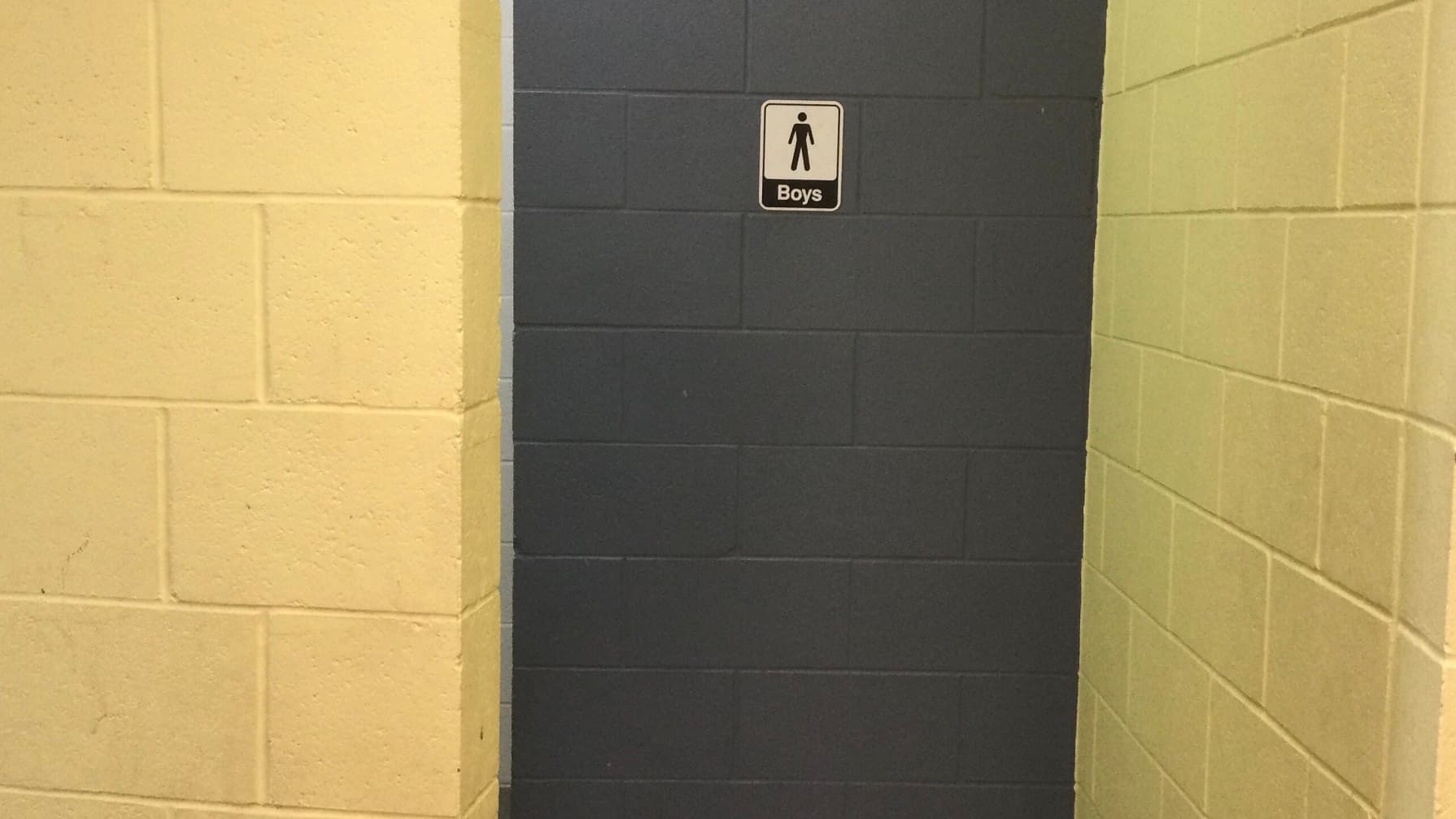 The male restroom's grey concrete wall has white and black signage with a male representation and the word “Boys”, while the yellow walls have a similar concrete design pattern.